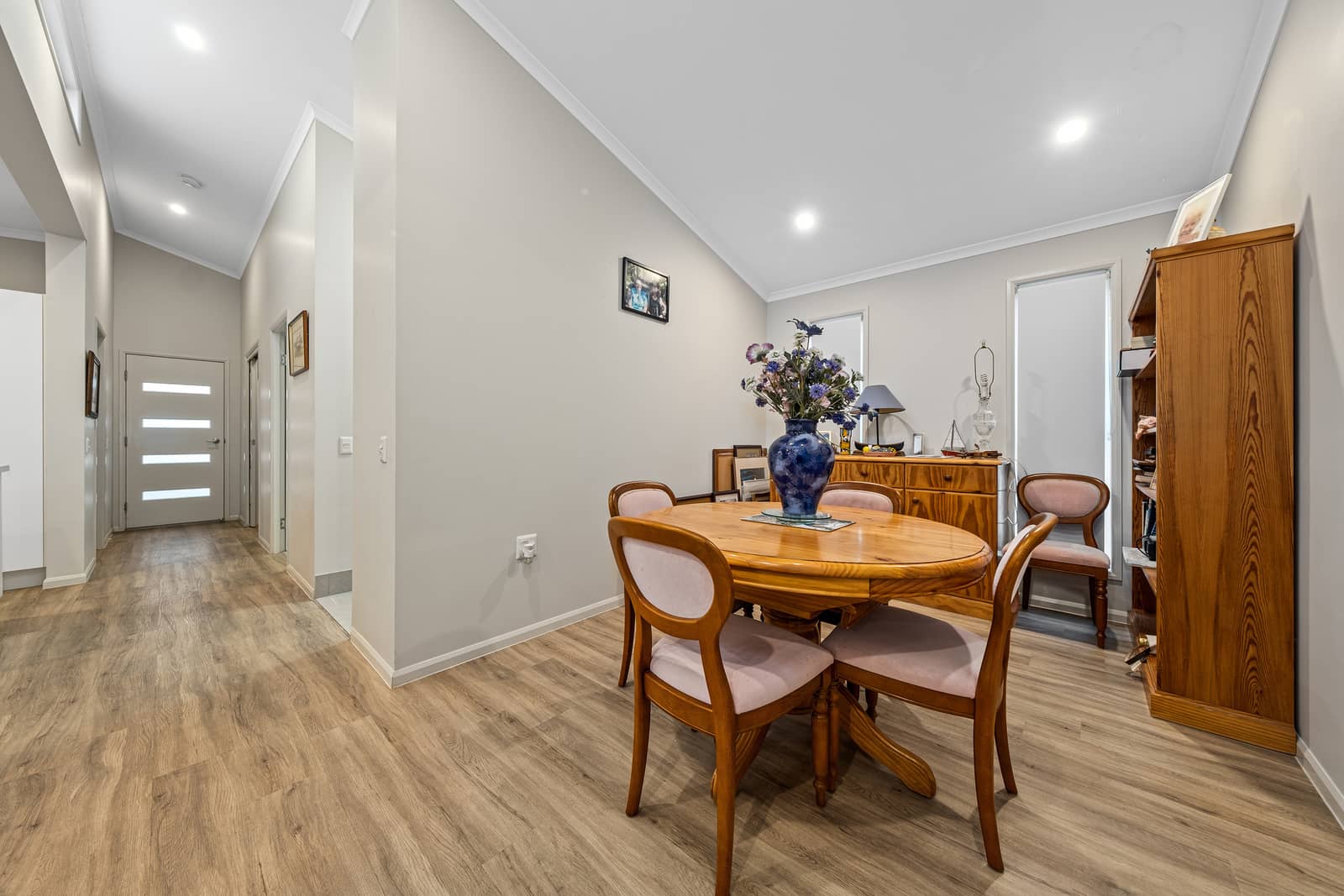 Dining area and hallway in an Ingenia Lifestyle home. Features a wooden oval table, upholstered chairs, wooden sideboard, and laminate flooring.