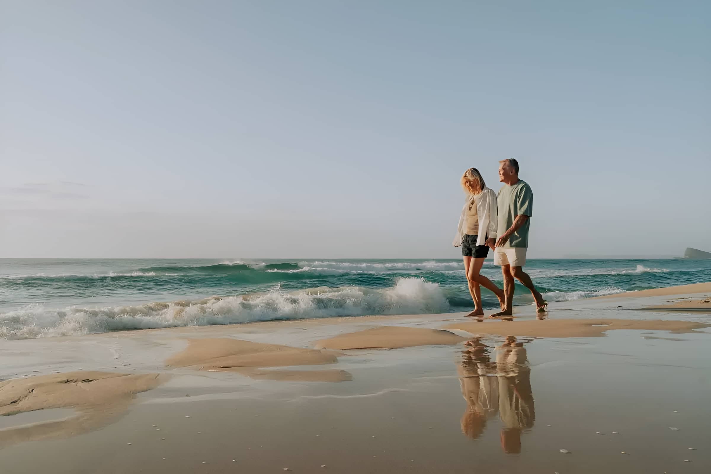 A woman and a man walk barefoot on a sandy beach at the ocean's edge, with waves breaking.