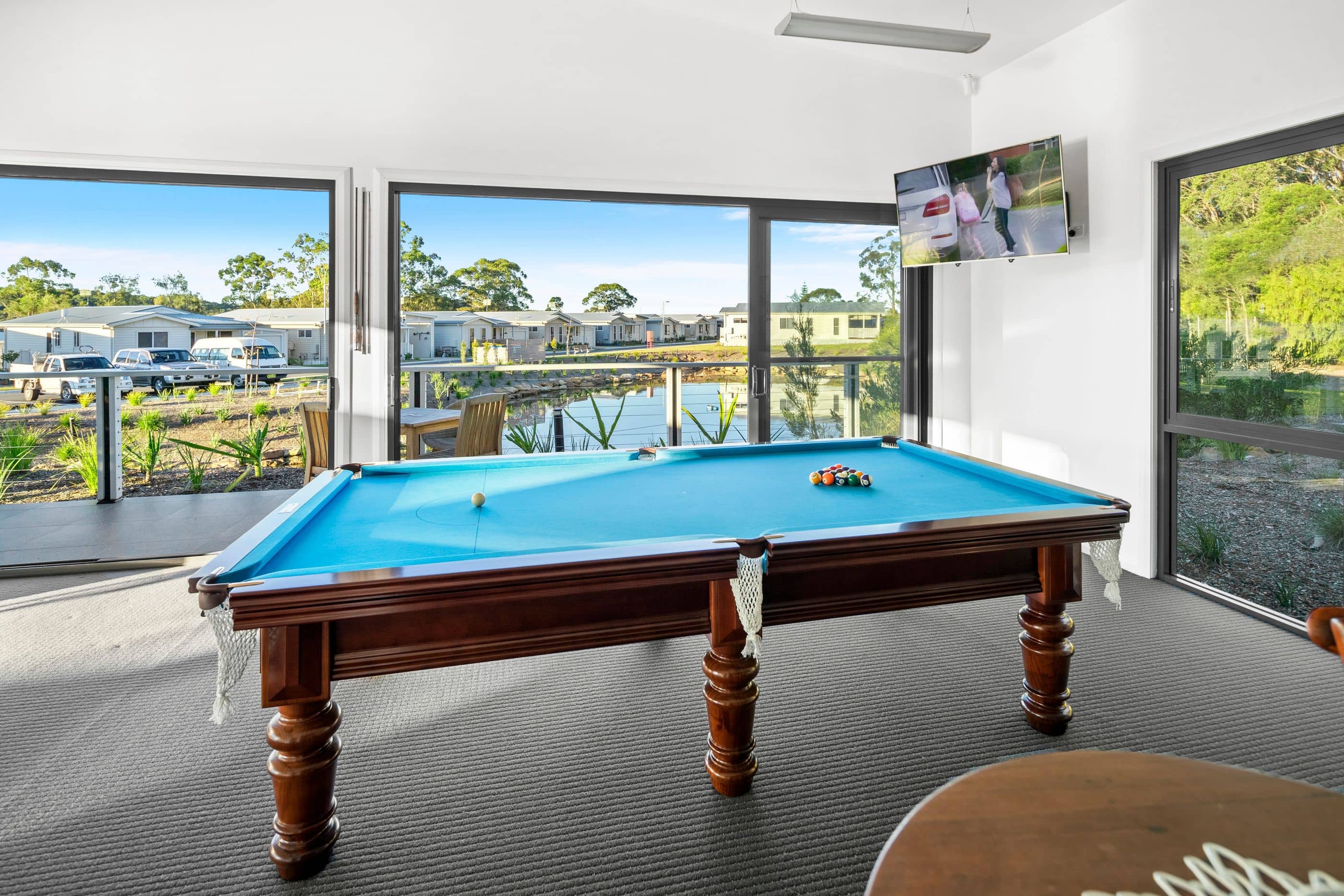 A blue billiards table is in the foreground, with pool balls racked and one ball on the table. Large windows show an Ingenia Lifestyle community with homes, a pond, and landscaped grounds.