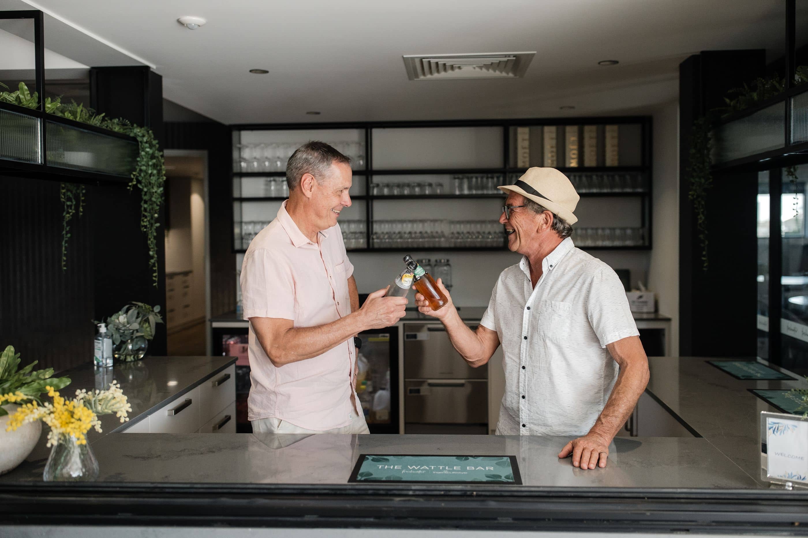 Two men smiling and clinking bottles at "The Wattle Bar" in an Ingenia Lifestyle community.