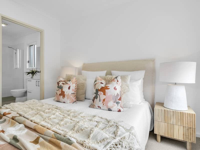 A neutral-toned bedroom with a bed, decorative pillows, and a bedside table with a lamp. A doorway shows a glimpse of an ensuite bathroom.