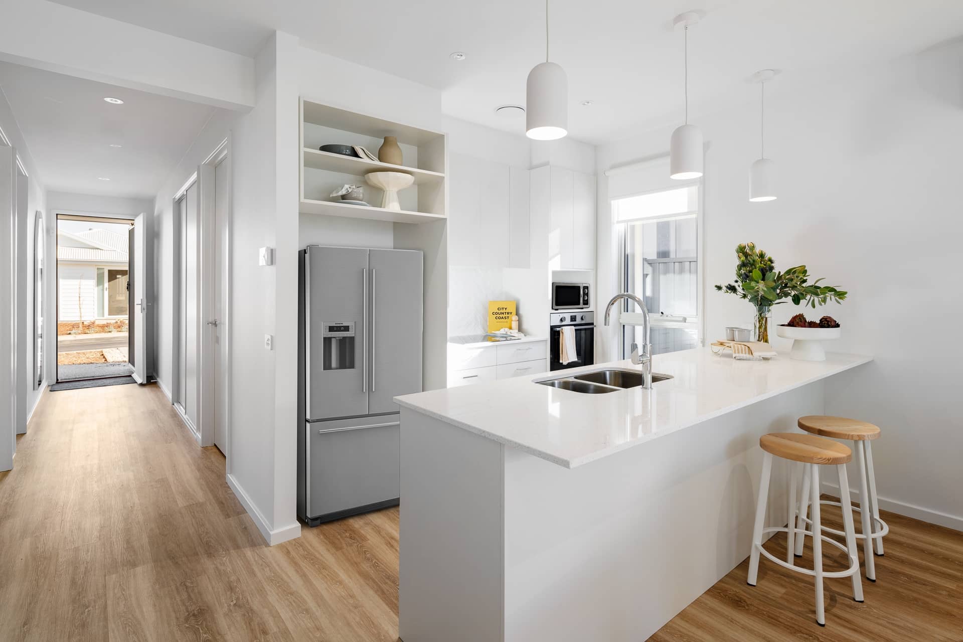 Modern kitchen of an Ingenia Lifestyle home featuring a white island, stainless steel appliances, and a hallway leading to a community street view.