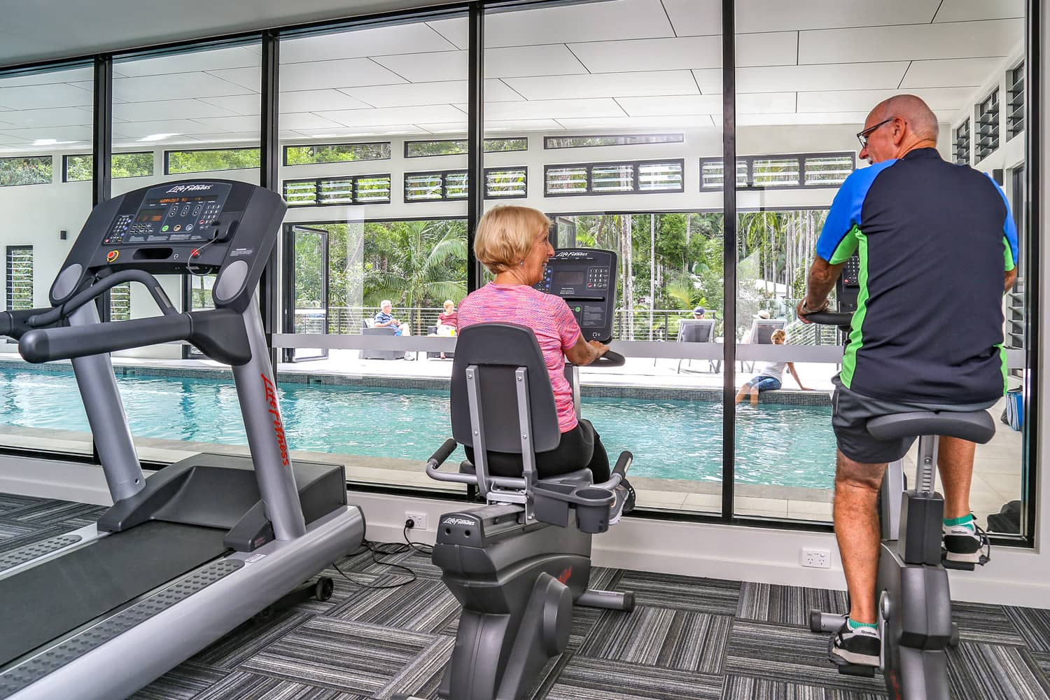 Two people use exercise bikes in a gym overlooking a swimming pool and lush greenery.