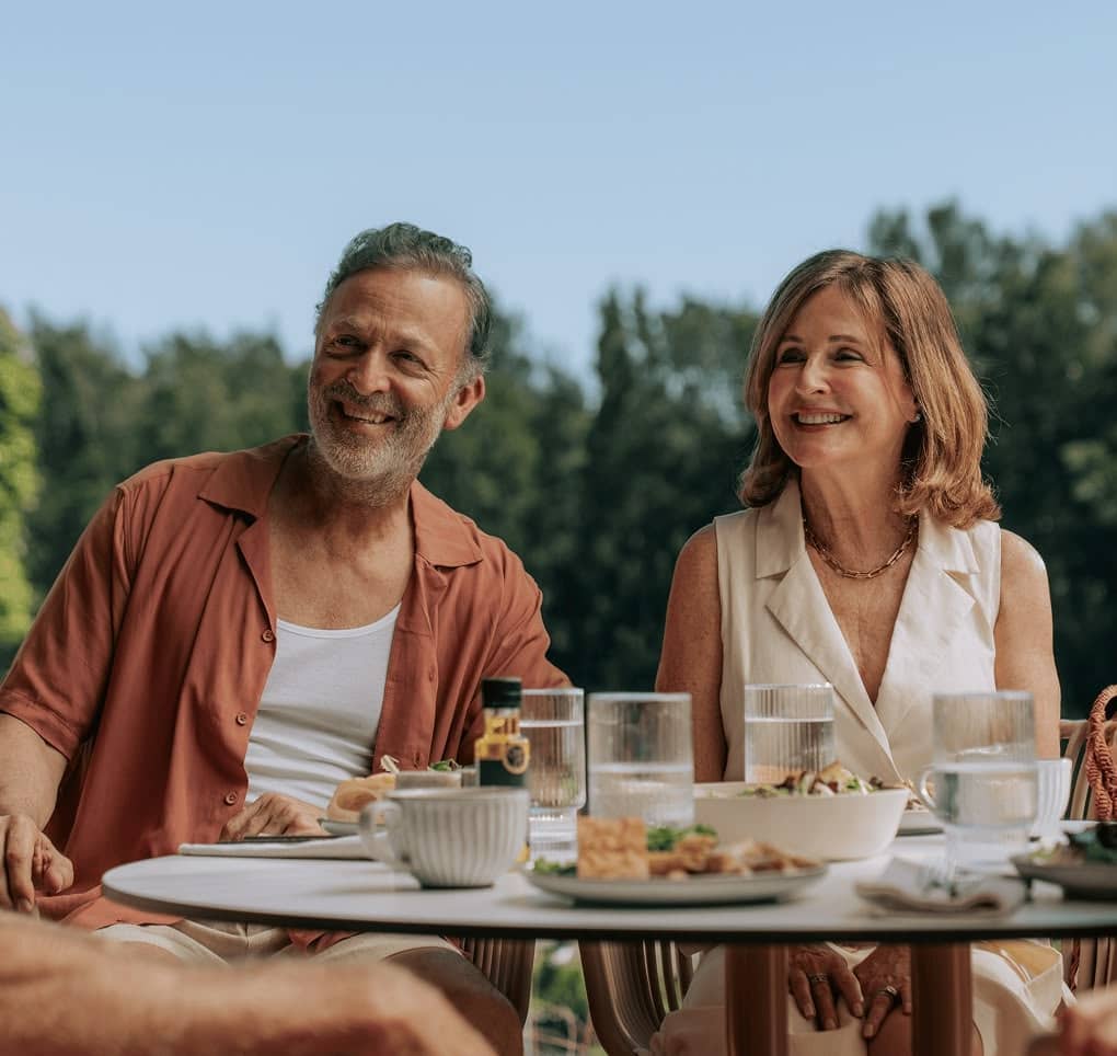 Two people smiling at an outdoor dining table with food and drinks. The scene suggests a relaxed meal in an Ingenia Lifestyle community.