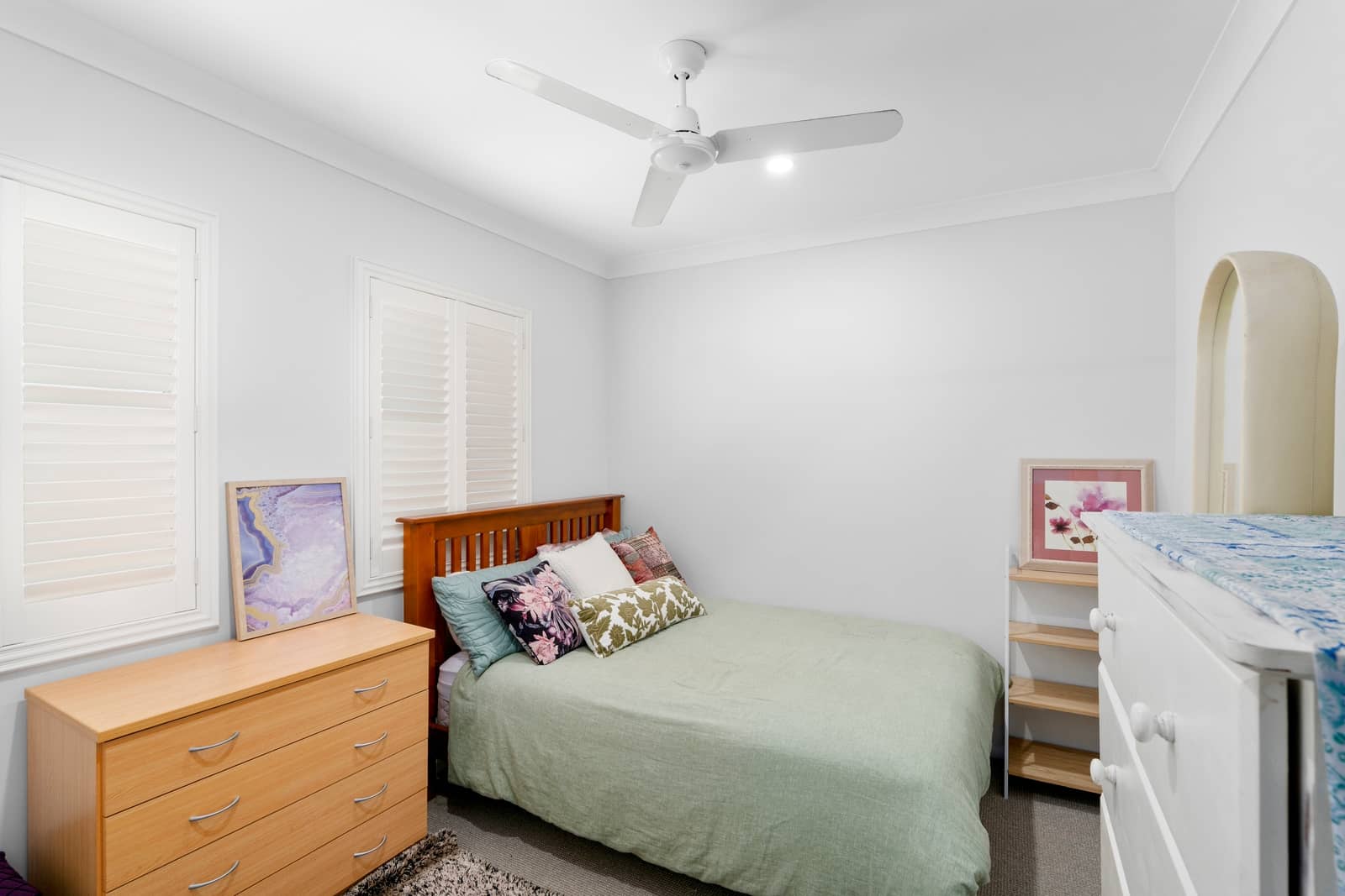 A bedroom with a double bed, light green duvet, and decorative pillows, next to a wooden dresser with artwork.