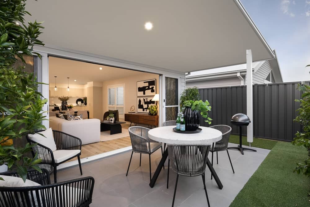 Outdoor entertaining area with a round table, chairs, and a BBQ, opening to a bright, modern living space.