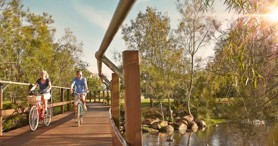 Two residents cycling on a wooden boardwalk next to a pond within an Ingenia Lifestyle community with lush trees.