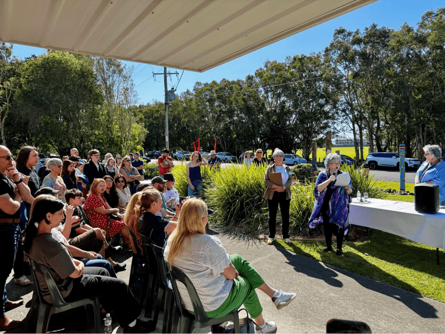 An outdoor community event under a canopy, with a speaker addressing a diverse group of attendees.