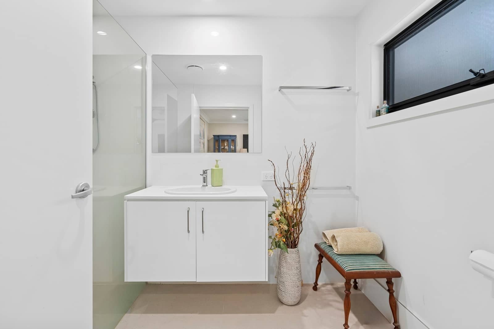 A bright bathroom featuring a white vanity with a sink and mirror, a glass-enclosed shower, and a small bench with towels.