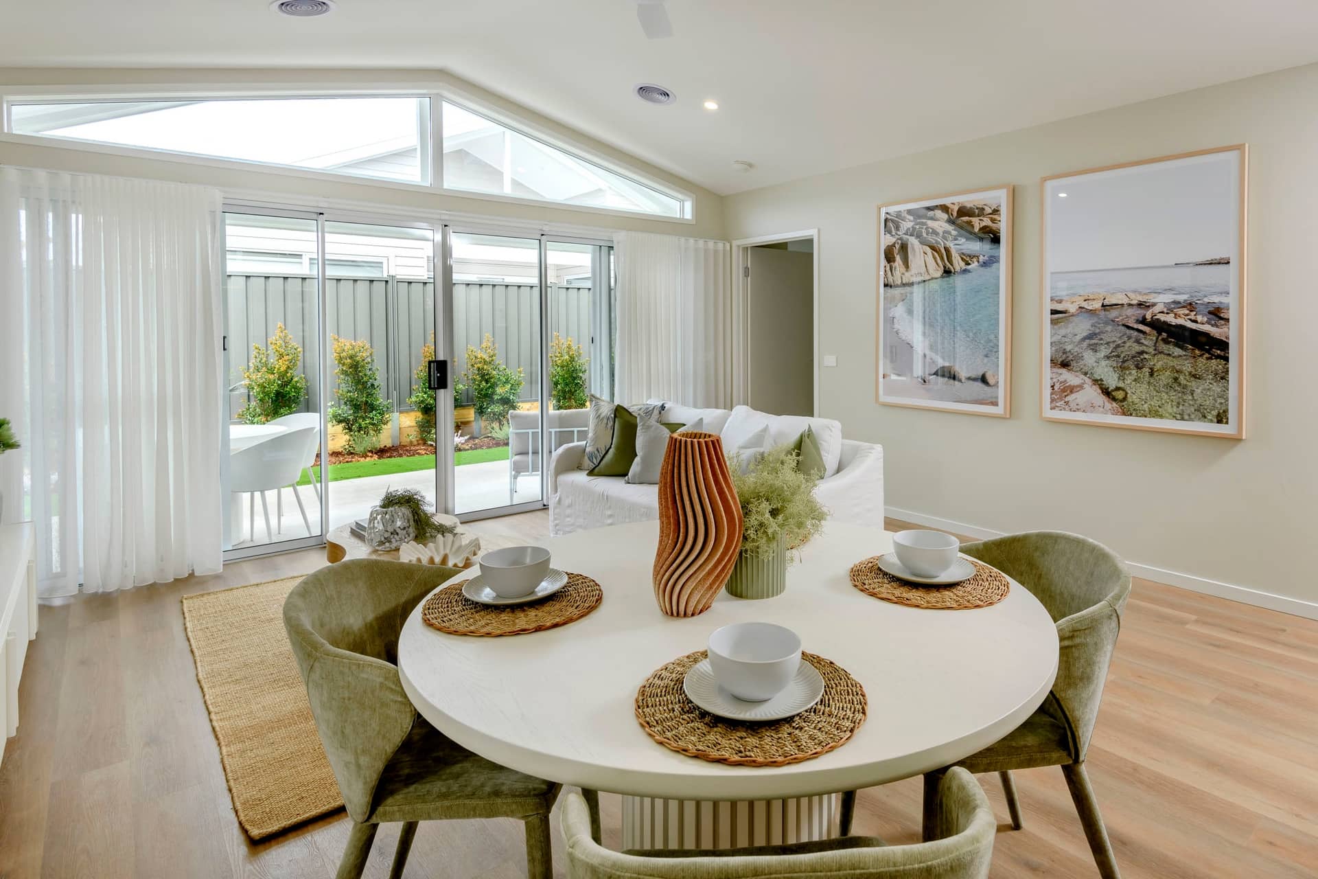 A circular white dining table with place settings is in the foreground of a living area with a sofa and large windows looking out to a garden.