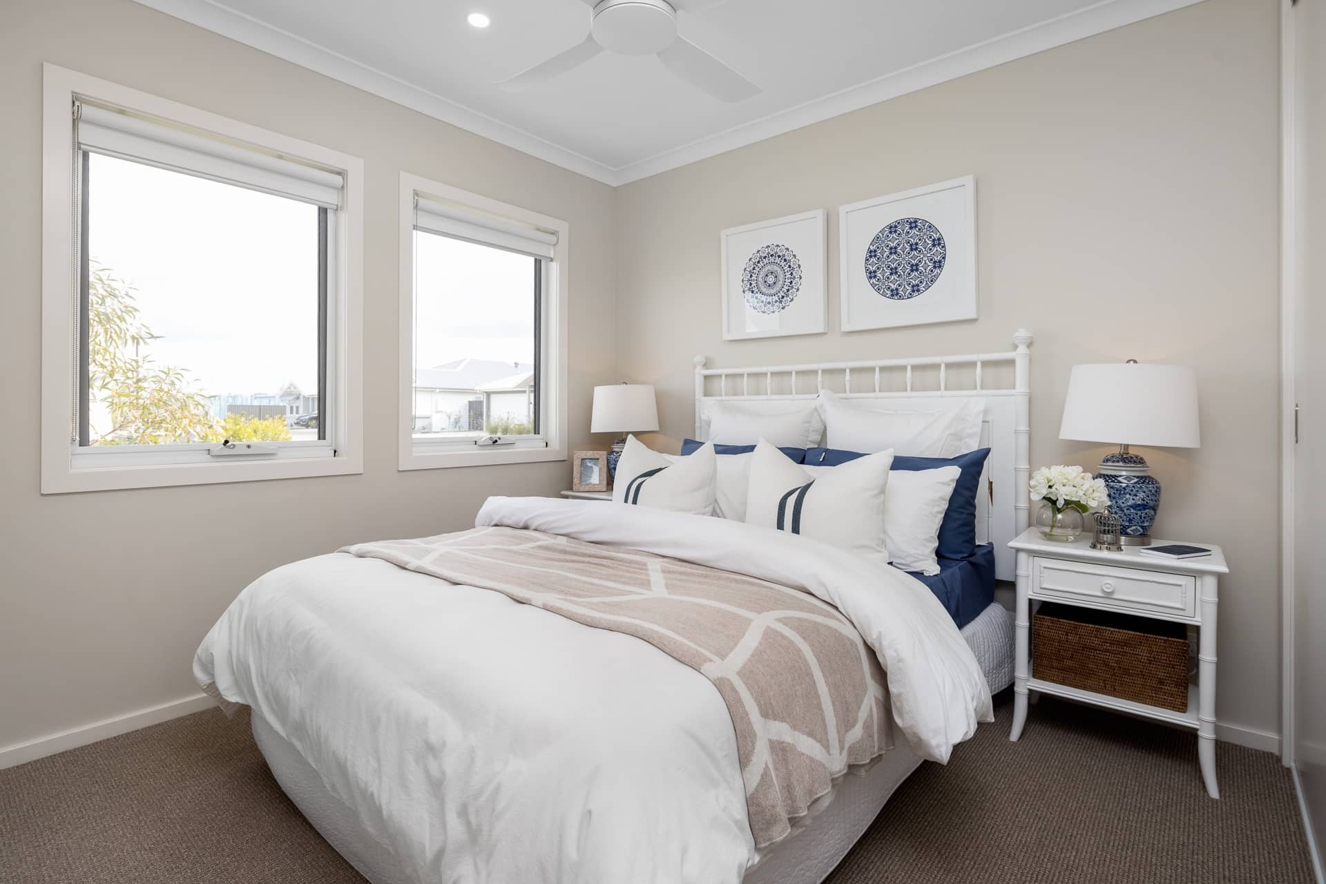 A neatly made bed with white bedding and a neutral throw in a bedroom with two windows and decorative wall art.