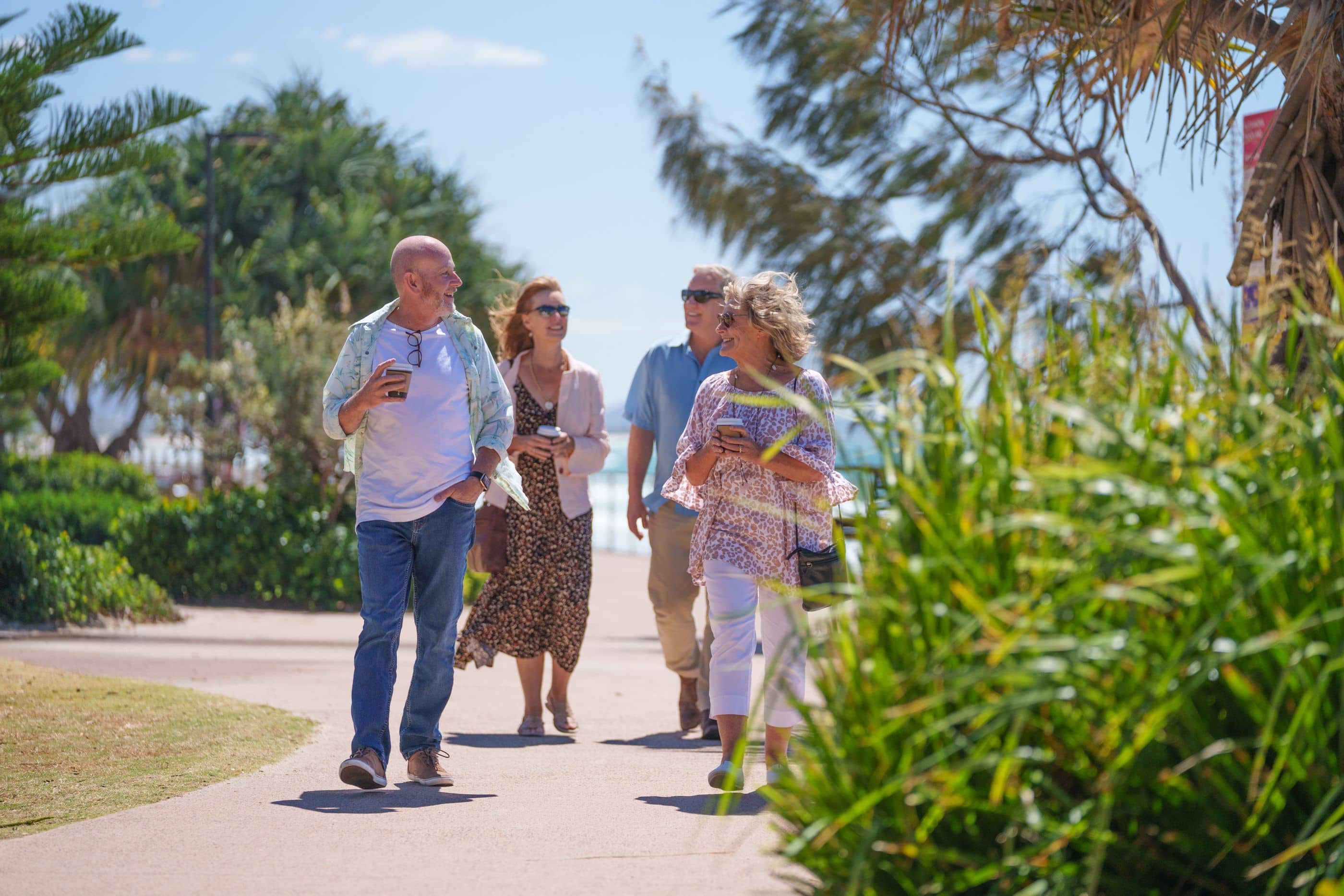 Four residents walk and chat on a sunny path in an Ingenia Lifestyle community, holding coffees.