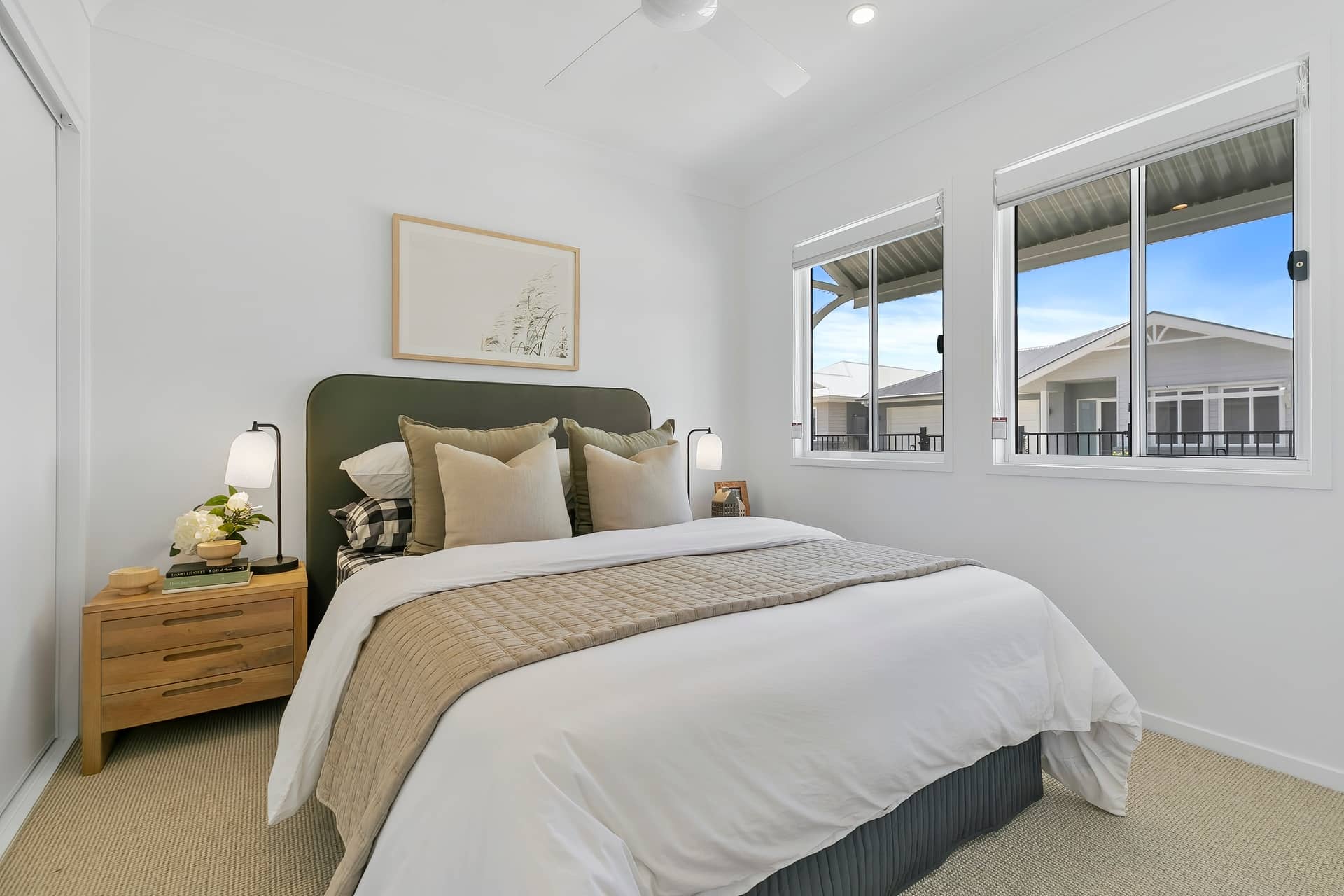 A bedroom with a neatly made bed, dark green headboard, wooden bedside table, and windows viewing an Ingenia Lifestyle community.