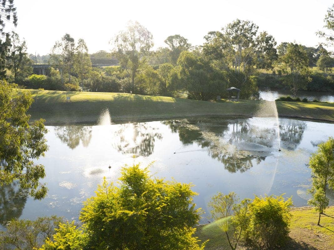 A pond with fountains and reflections of trees, bordered by manicured lawns and lush greenery.