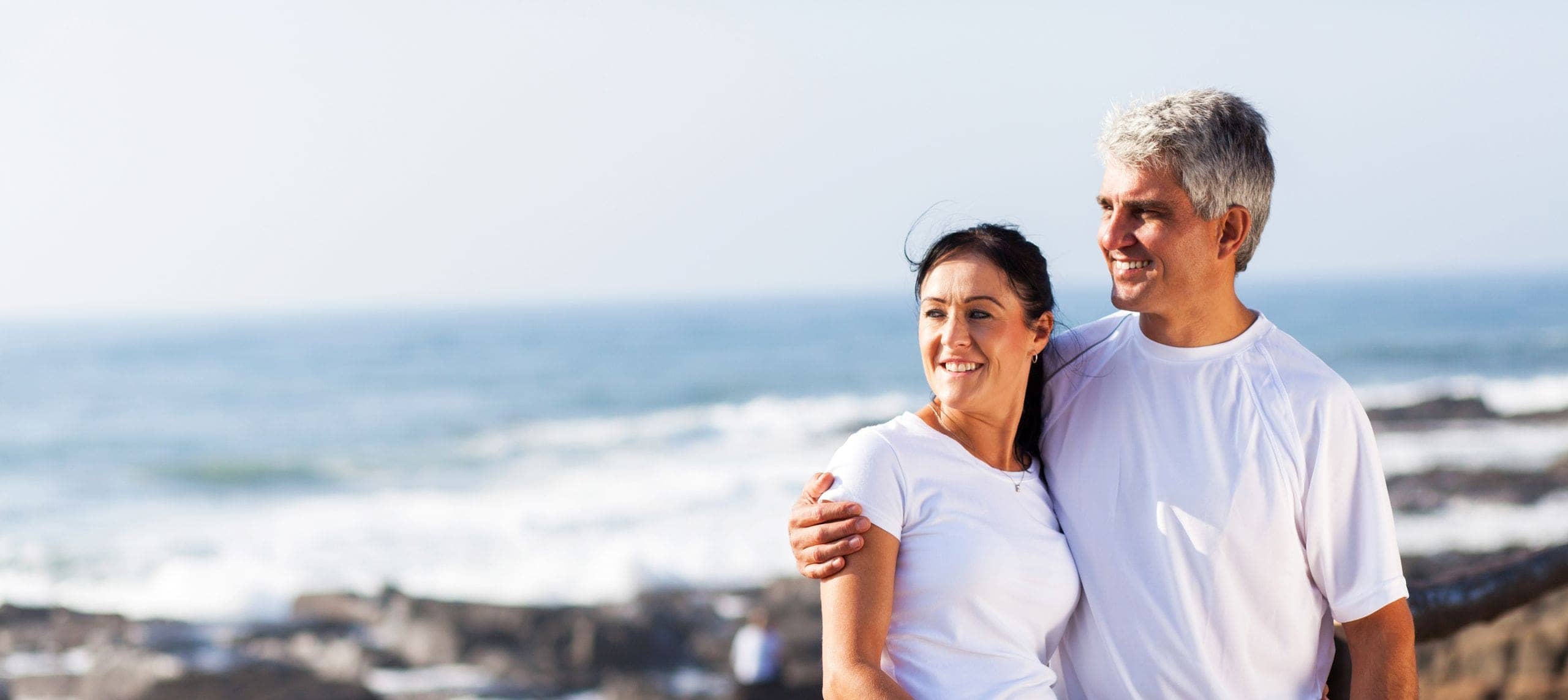 Smiling man and woman by the ocean, with waves and rocks in the background.