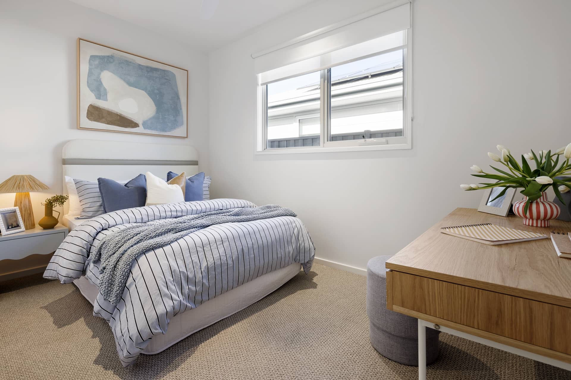 A bedroom with a bed, blue and white striped bedding, a wooden desk, and a window. Beige carpet and white walls, suitable for an Ingenia Lifestyle home.