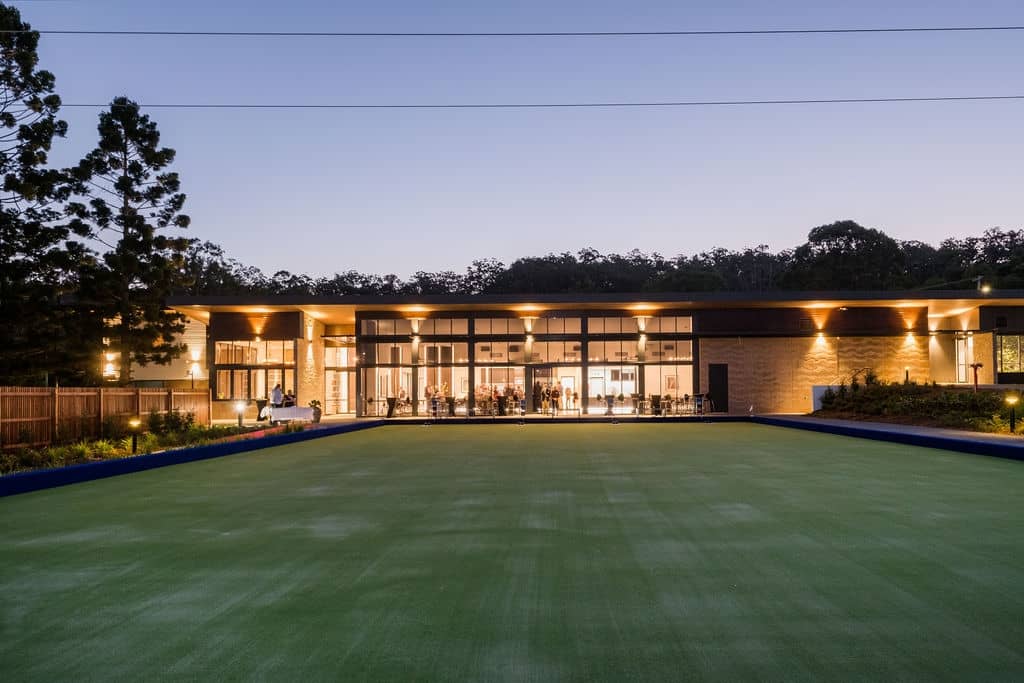 A green bowling green in front of a modern community centre with large windows and outdoor lighting.