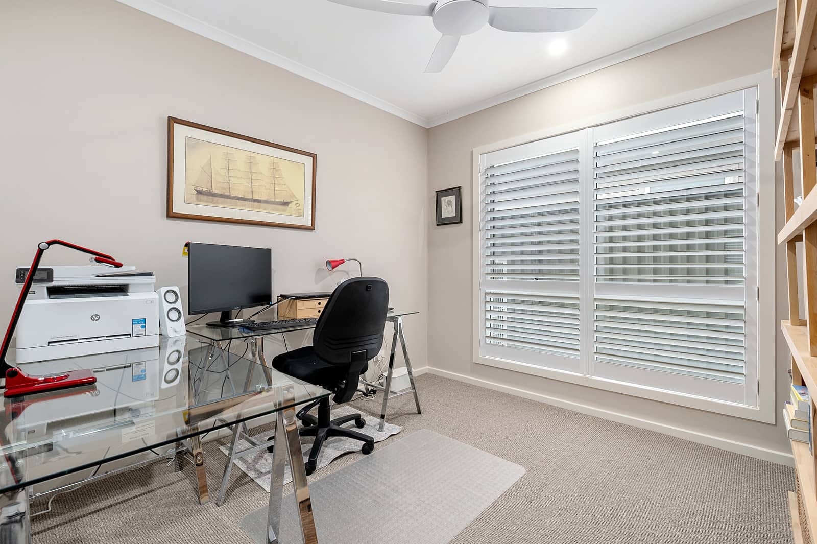 A modern home office with a glass desk, computer, and a large window with white plantation shutters in an over-55 home.