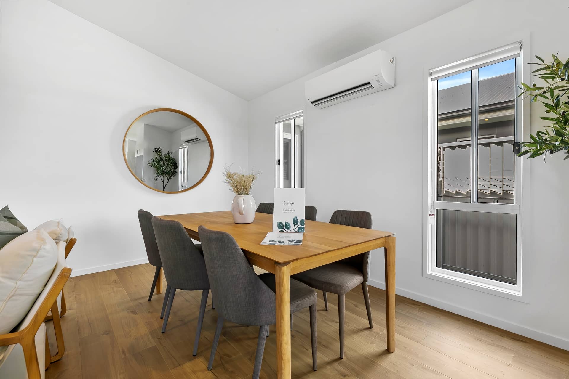 Modern dining area of an Ingenia Lifestyle home with a wooden table, grey chairs, wall mirror, and air conditioning.