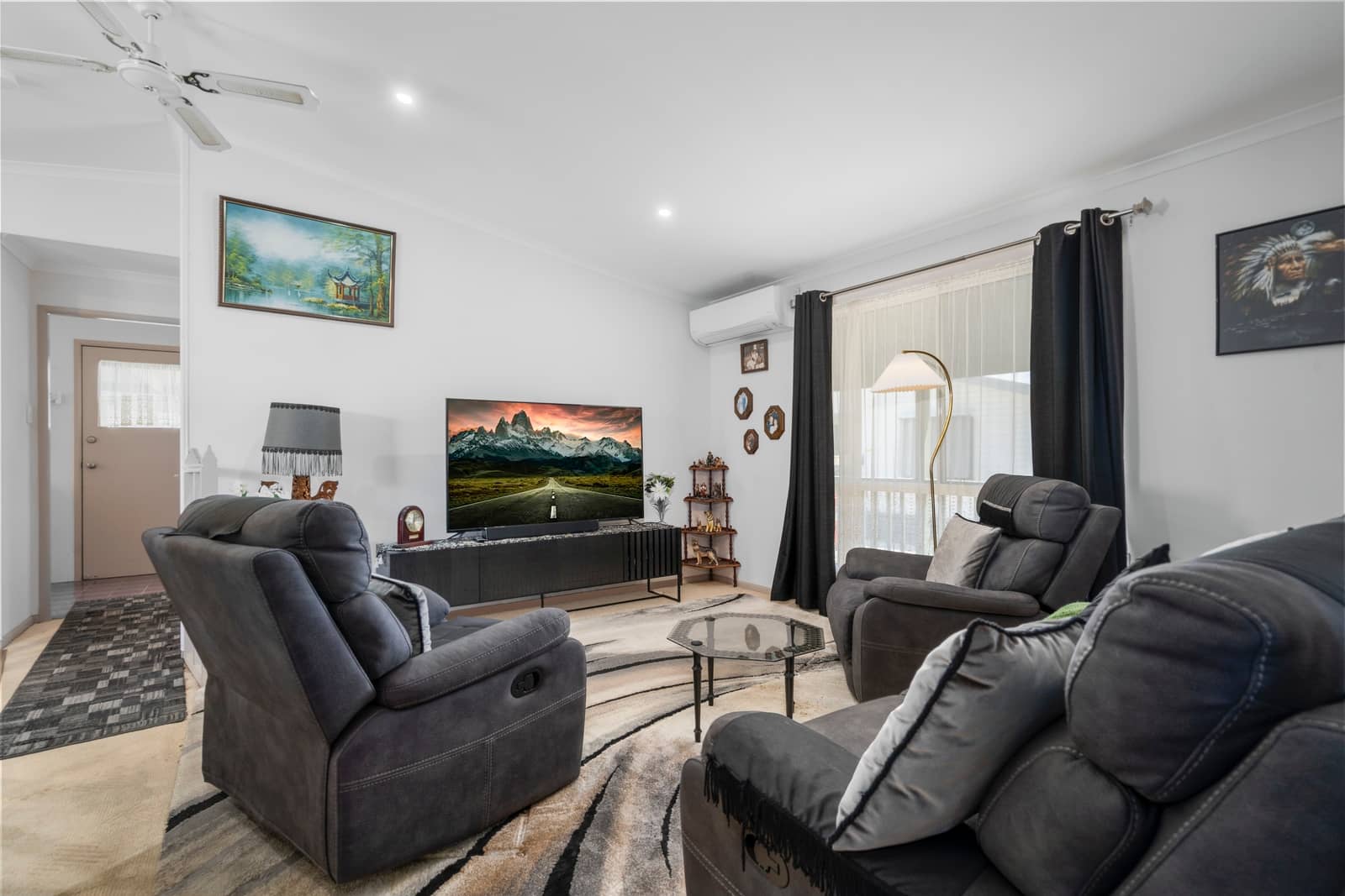 A furnished living room in an Ingenia Lifestyle home featuring grey recliners, a large TV, and a patterned area rug.