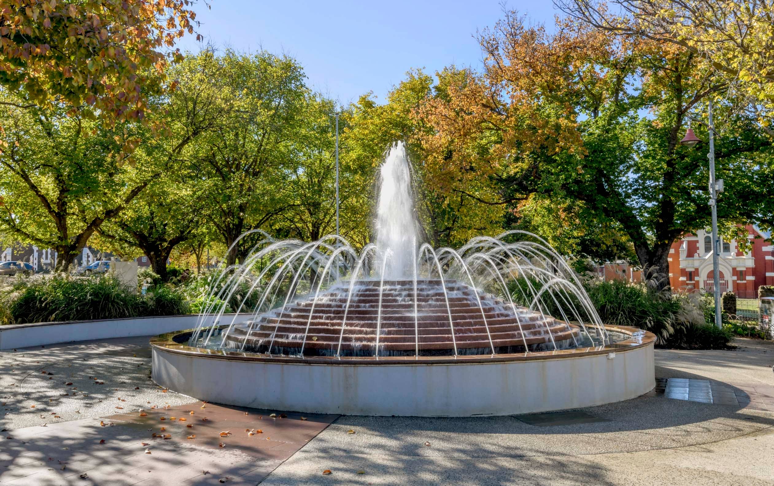 Tiered fountain with water jets, surrounded by trees with autumn foliage and manicured landscaping.