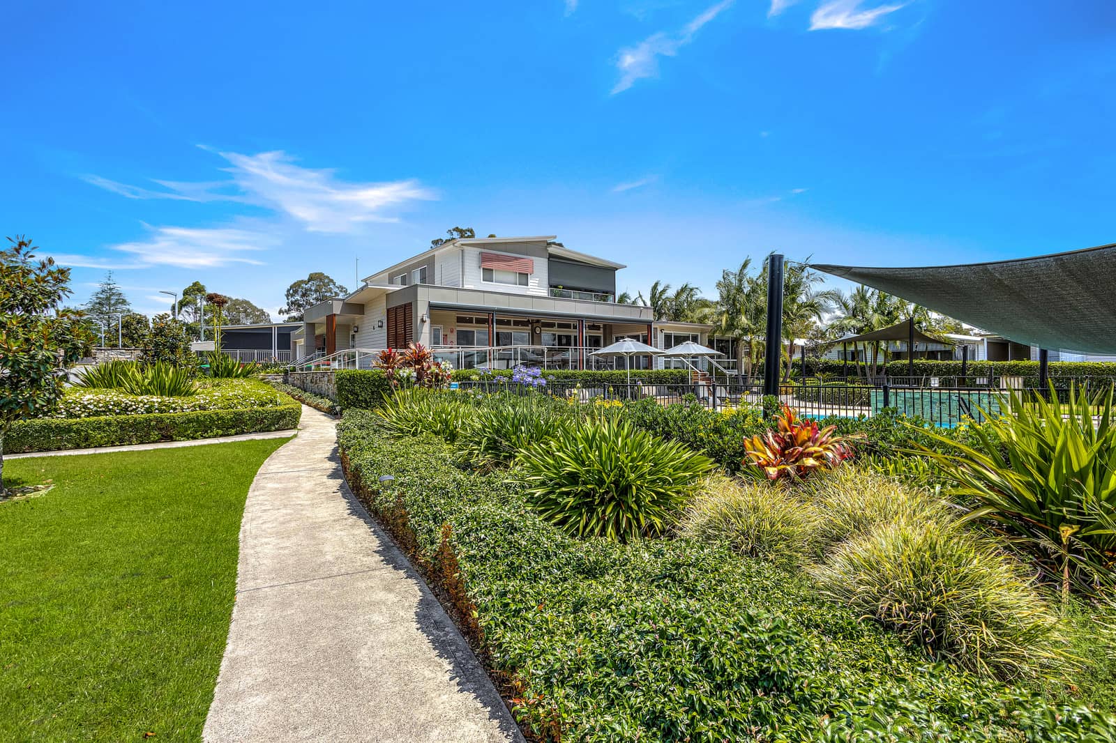 A paved path winds through landscaped gardens towards a modern building with a pool and shaded seating area.