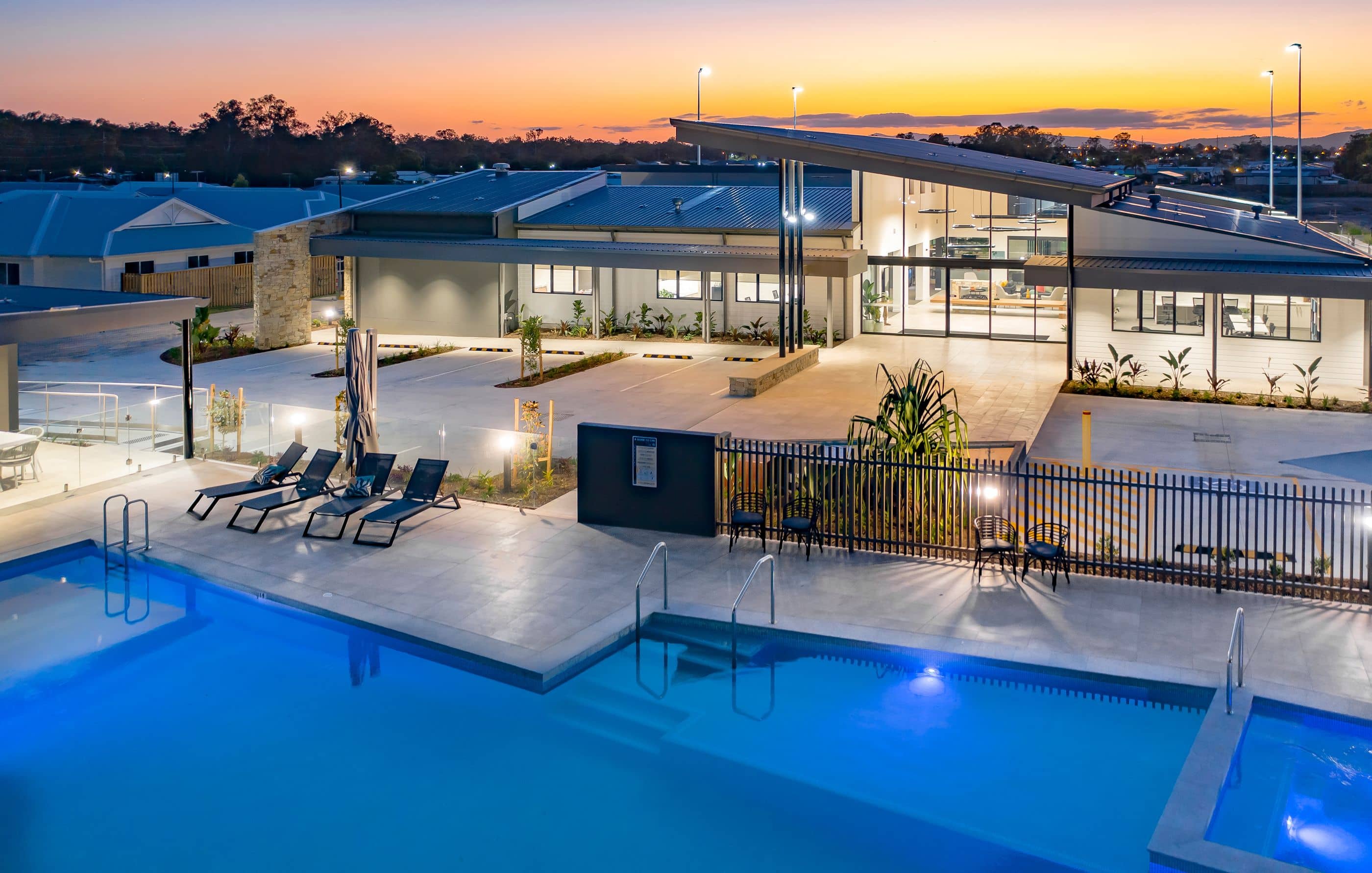 An Ingenia Lifestyle community's modern clubhouse and illuminated shared swimming pool at sunset, with lounge chairs.