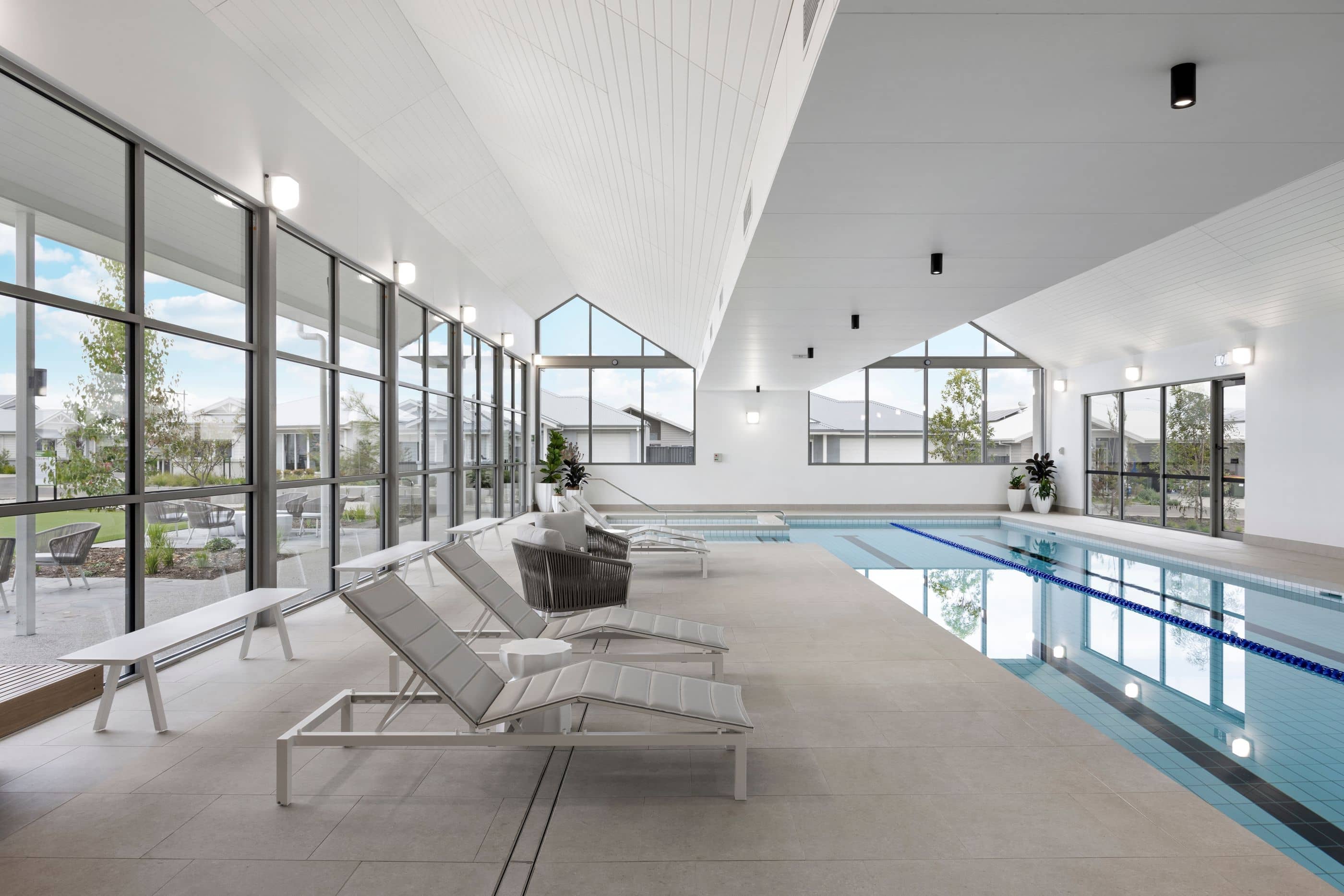 Bright indoor swimming pool at an Ingenia Lifestyle community, with lounge chairs, benches, and large windows revealing landscaped grounds.