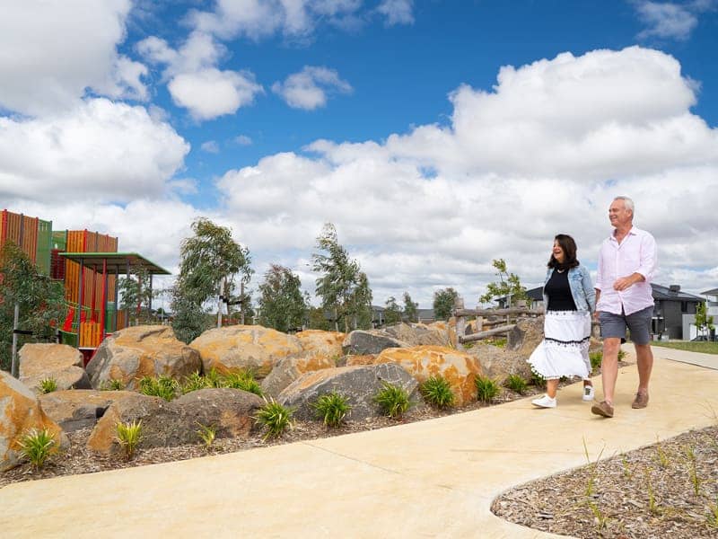 Two people walk on a paved path past landscaped rocks, trees, and a colourful playground within an Ingenia Lifestyle community.