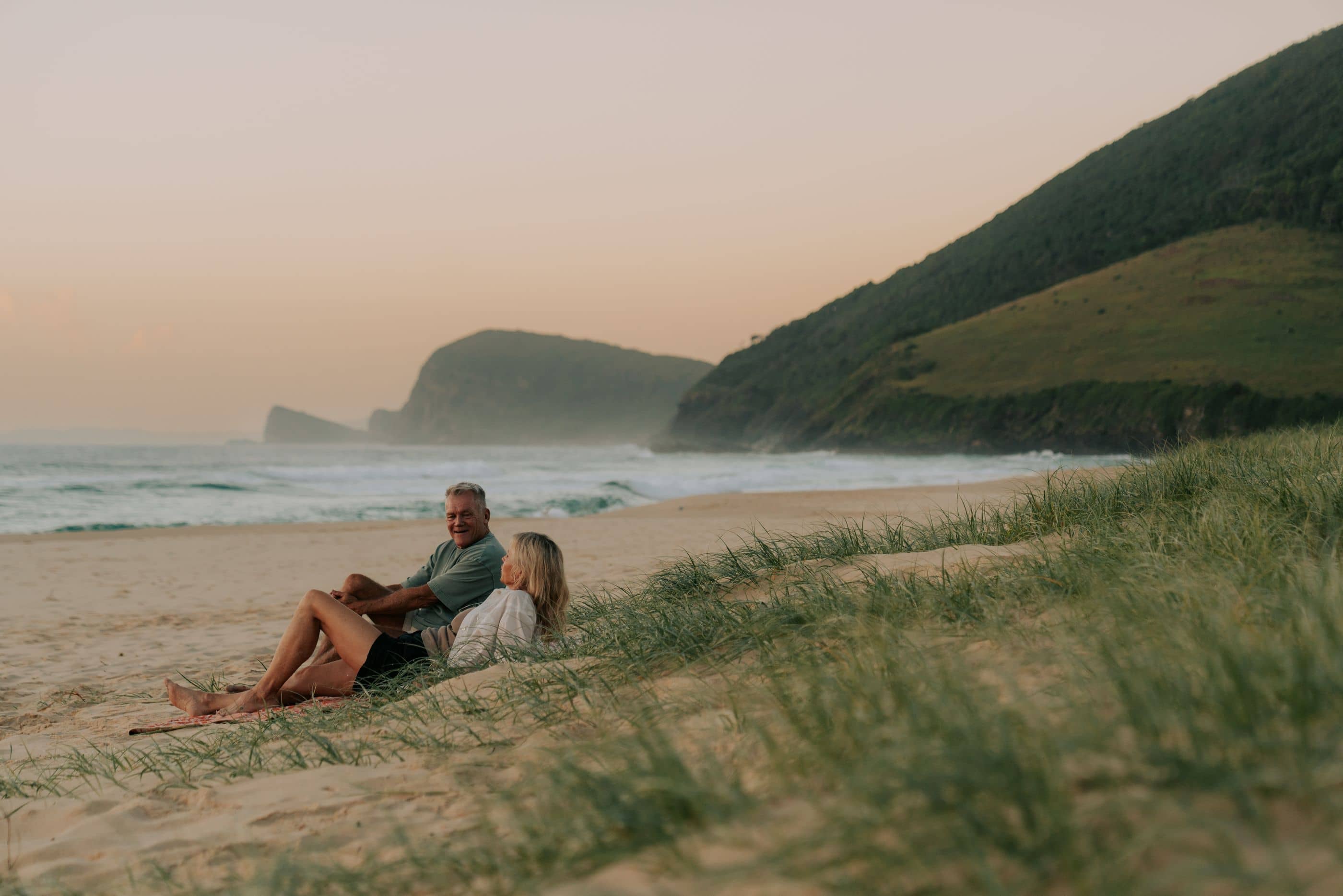 A man and a woman relax on a beach with waves in the background and green hills beyond.
