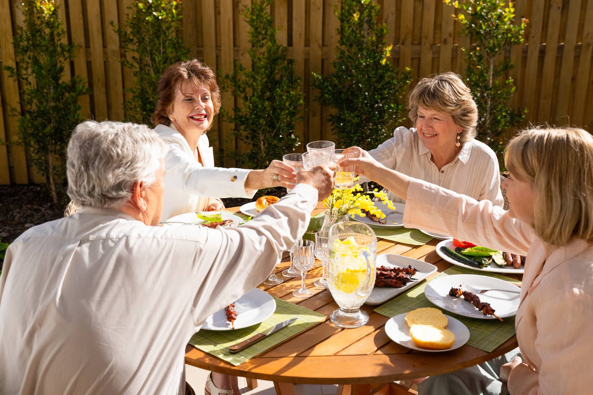 Four people enjoy a meal and toast with drinks at a wooden table in an Ingenia Lifestyle community garden.