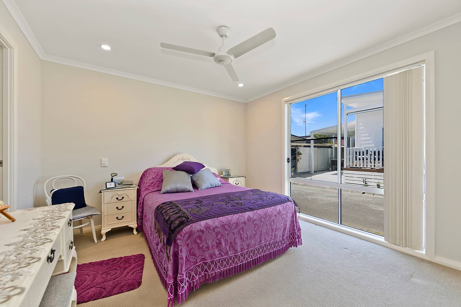 Bright bedroom with purple and grey bed, white furniture, ceiling fan, and large window viewing an Ingenia Lifestyle community.