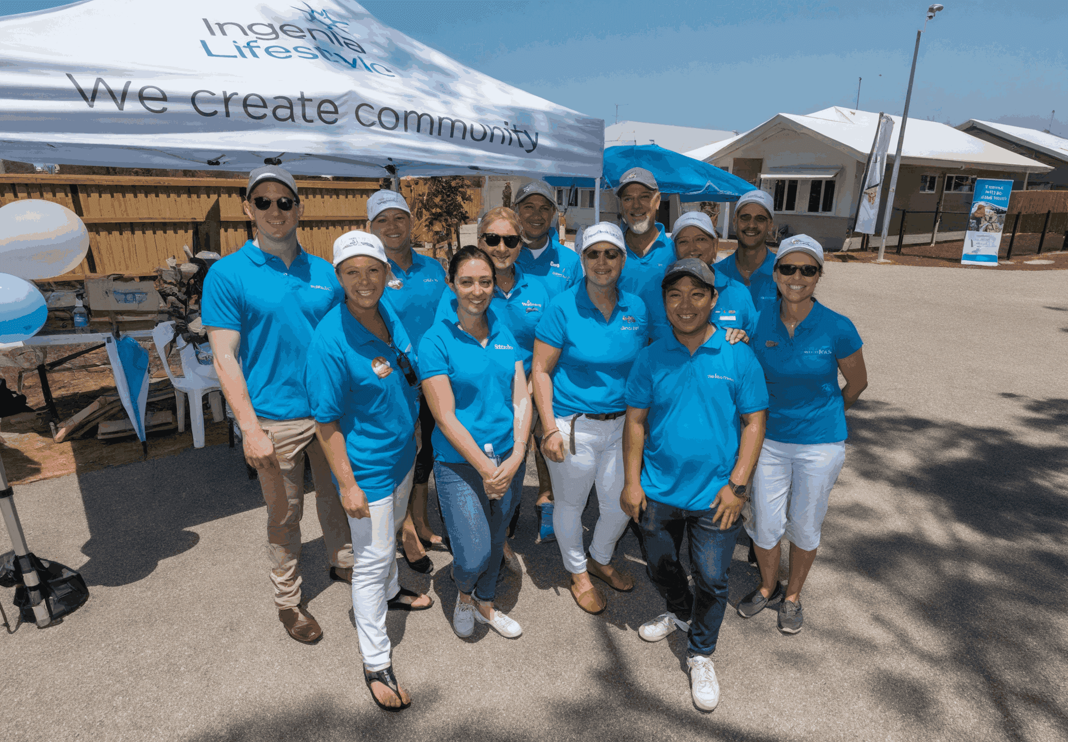 Twelve Ingenia Lifestyle team members in blue shirts and hats stand by a 'We create community' tent with buildings behind.