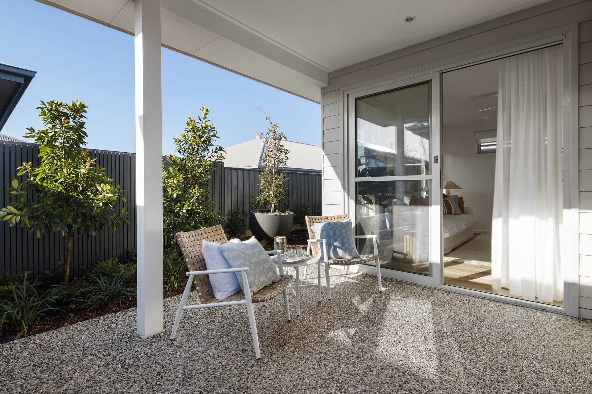 Covered alfresco patio of an Ingenia Lifestyle home with two chairs, a table, and low-maintenance landscaping. Sliding doors to the living room.