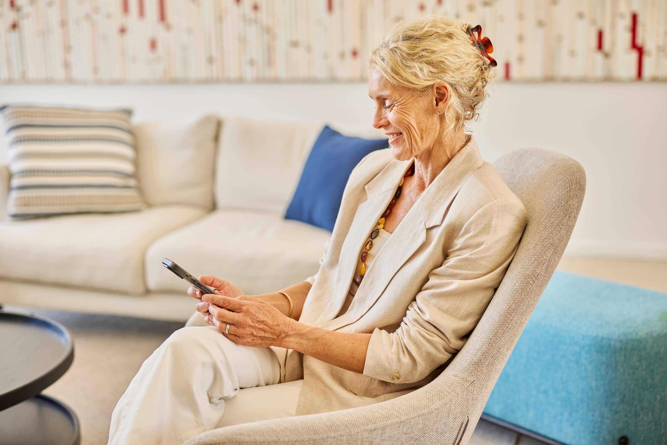 Smiling resident looking at her phone, seated in an armchair in a modern Ingenia Lifestyle community lounge.
