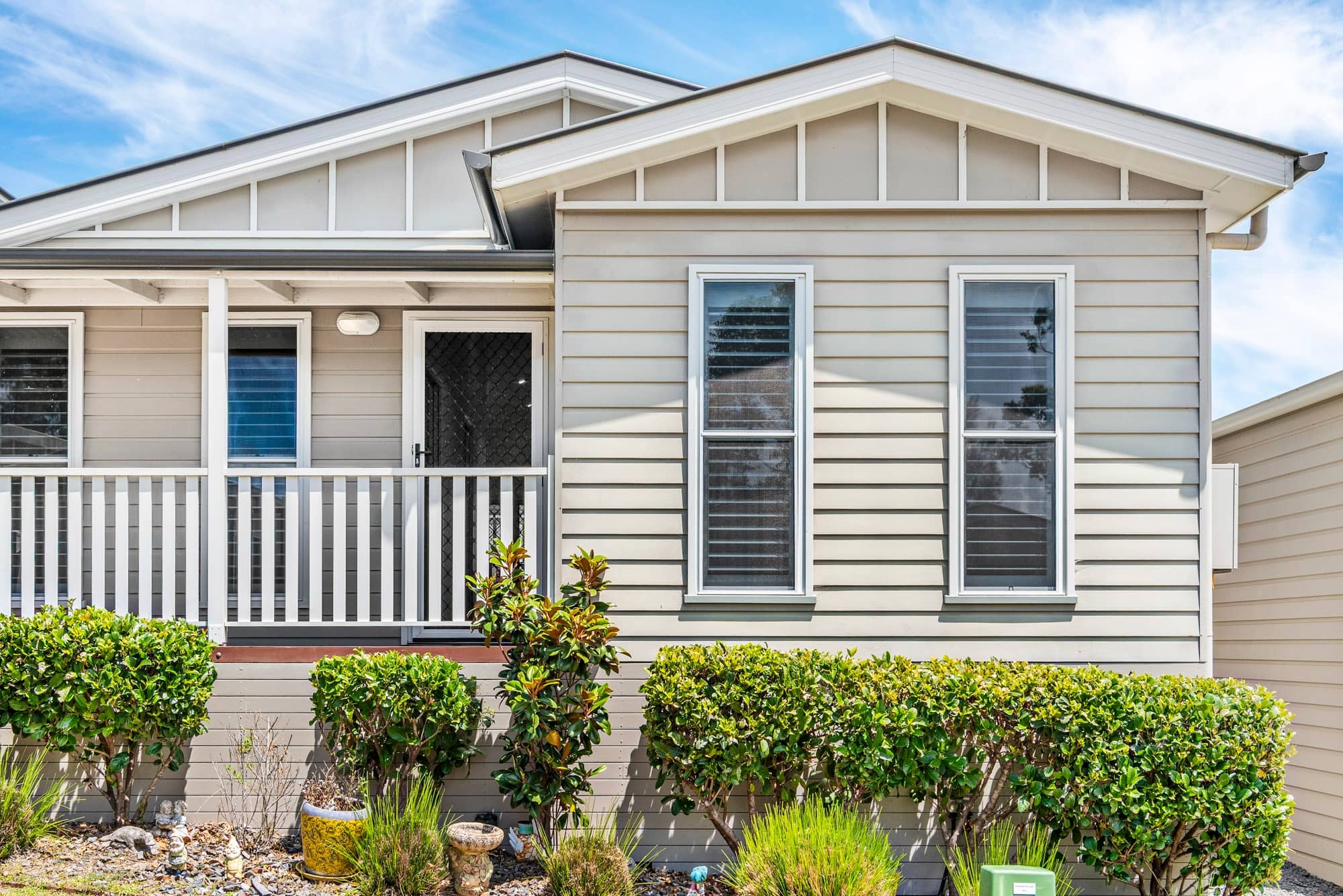 Front exterior of a modern, light beige land lease home with a porch, white railing, and low-maintenance landscaping under a blue sky.