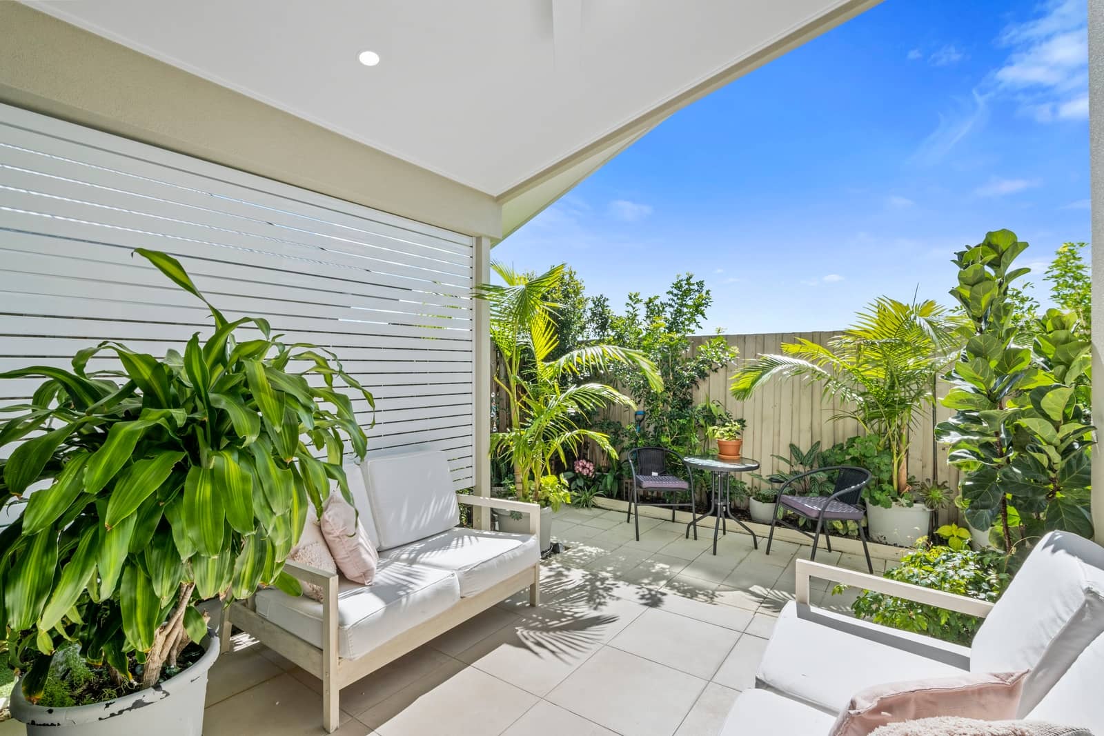 A covered outdoor patio at an Ingenia Lifestyle home, featuring white lounge chairs, lush greenery, and a small dining area.