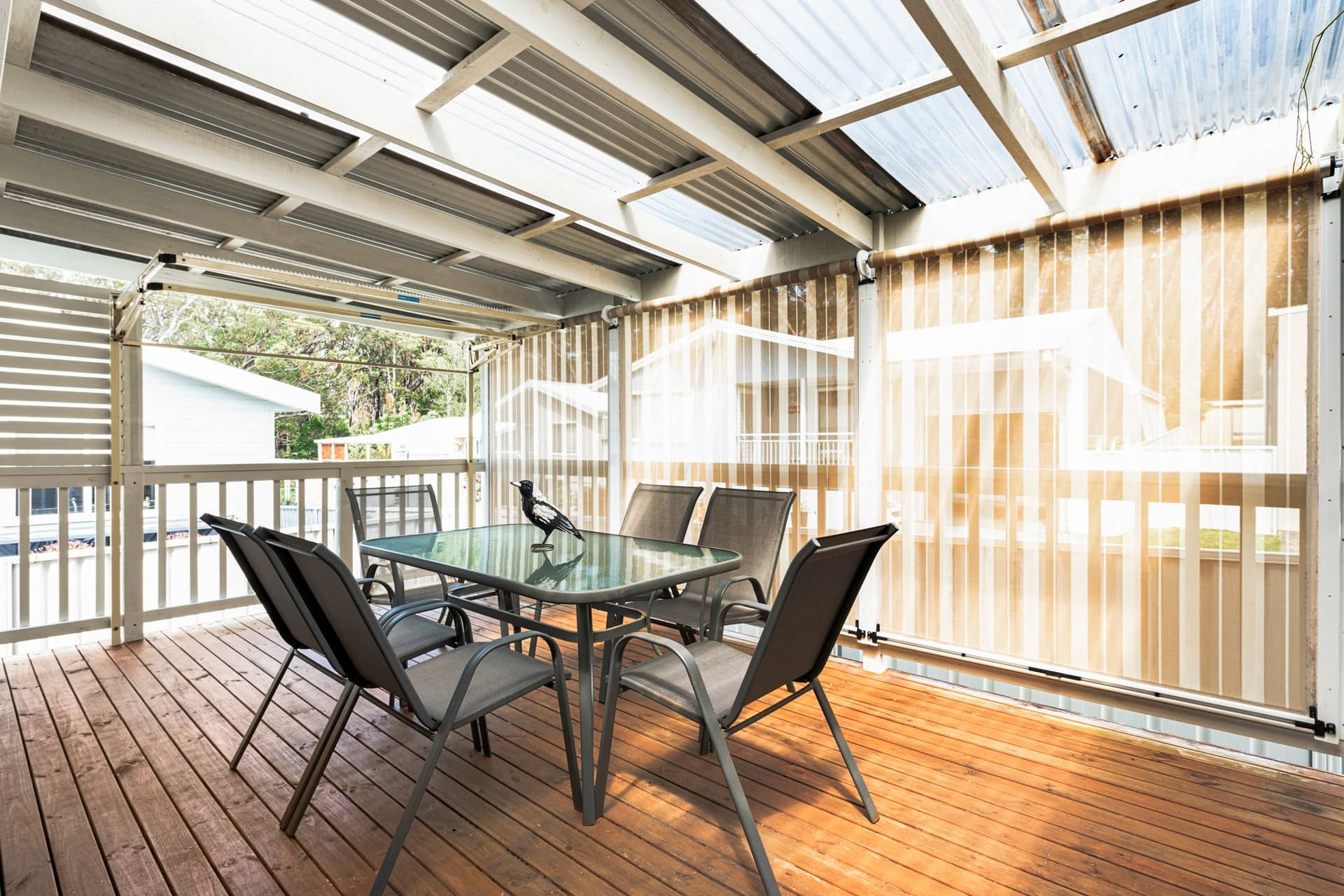 A covered wooden deck with a dining table, chairs, privacy screens, and a magpie. Other homes are visible in an Ingenia Lifestyle community.