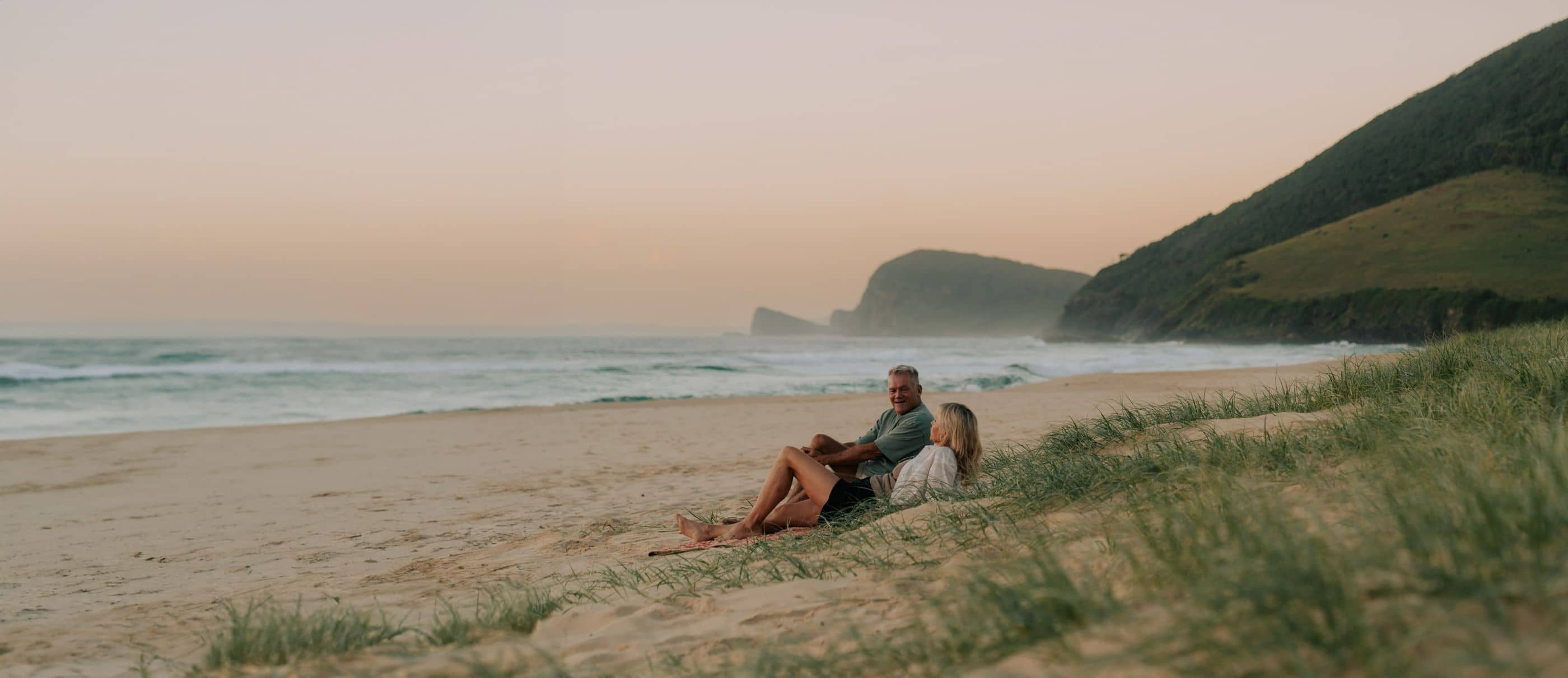 Two people sit on a sandy beach next to dunes with ocean and green hills in the background.