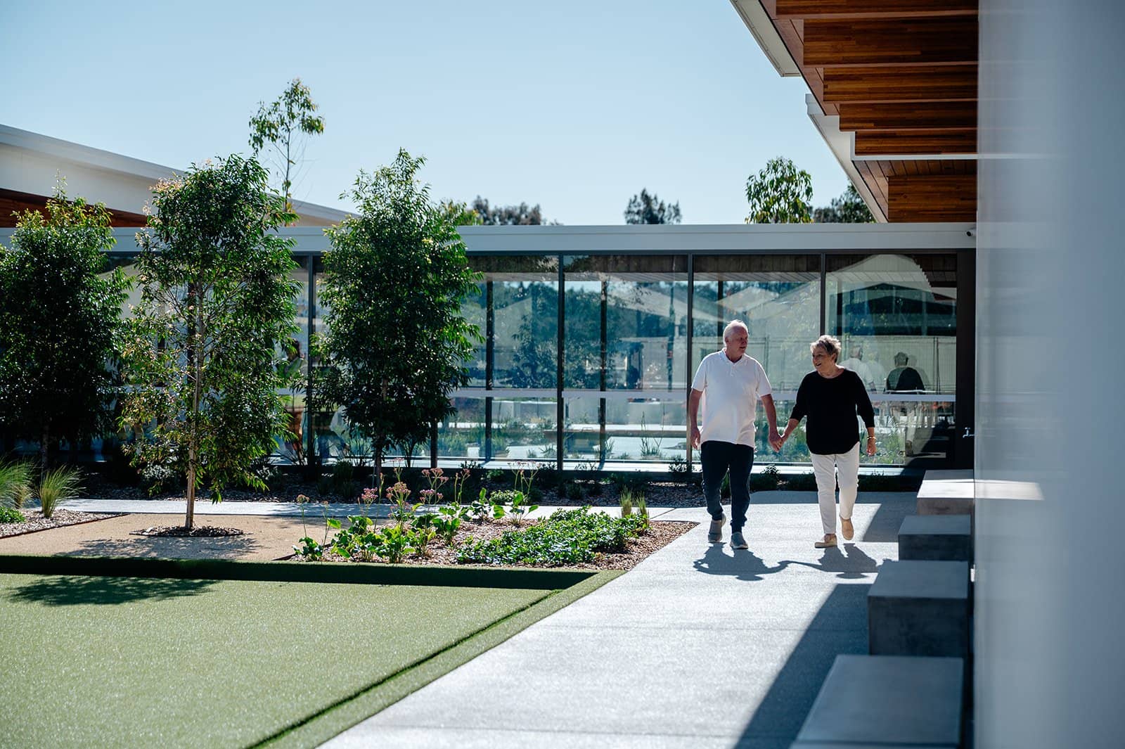 A couple walking hand-in-hand on a path in an Ingenia Lifestyle community with modern architecture and landscaped grounds.