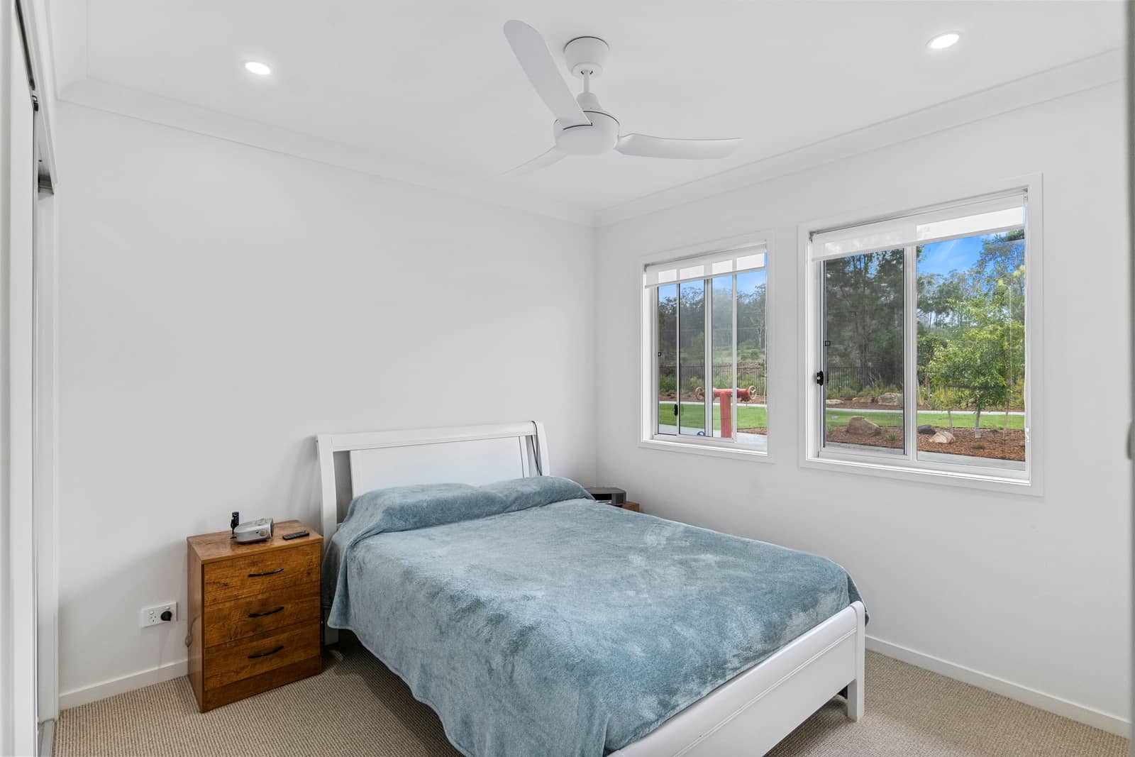 Bright bedroom in an Ingenia Lifestyle over-55 home, with a white bed, wooden bedside table, and windows with garden views.