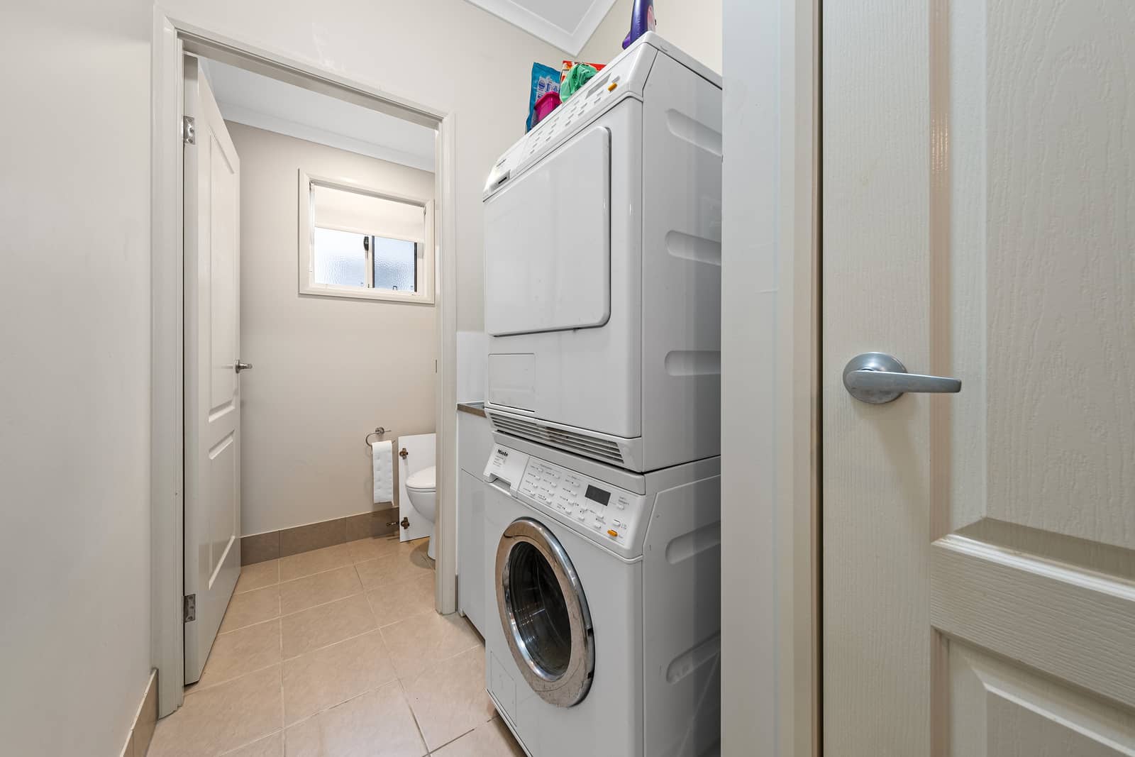 Stacked washing machine and dryer in a laundry space, with a compact bathroom visible beyond in an Ingenia Lifestyle home.
