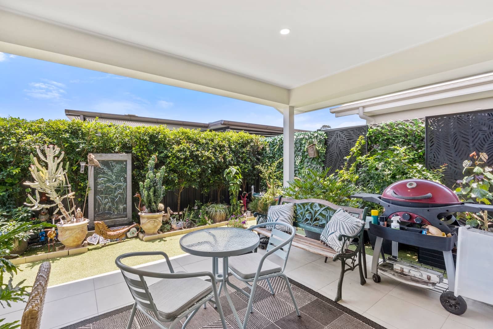 Covered alfresco patio with outdoor dining set and BBQ, overlooking a low-maintenance garden with hedges and decorative plants.