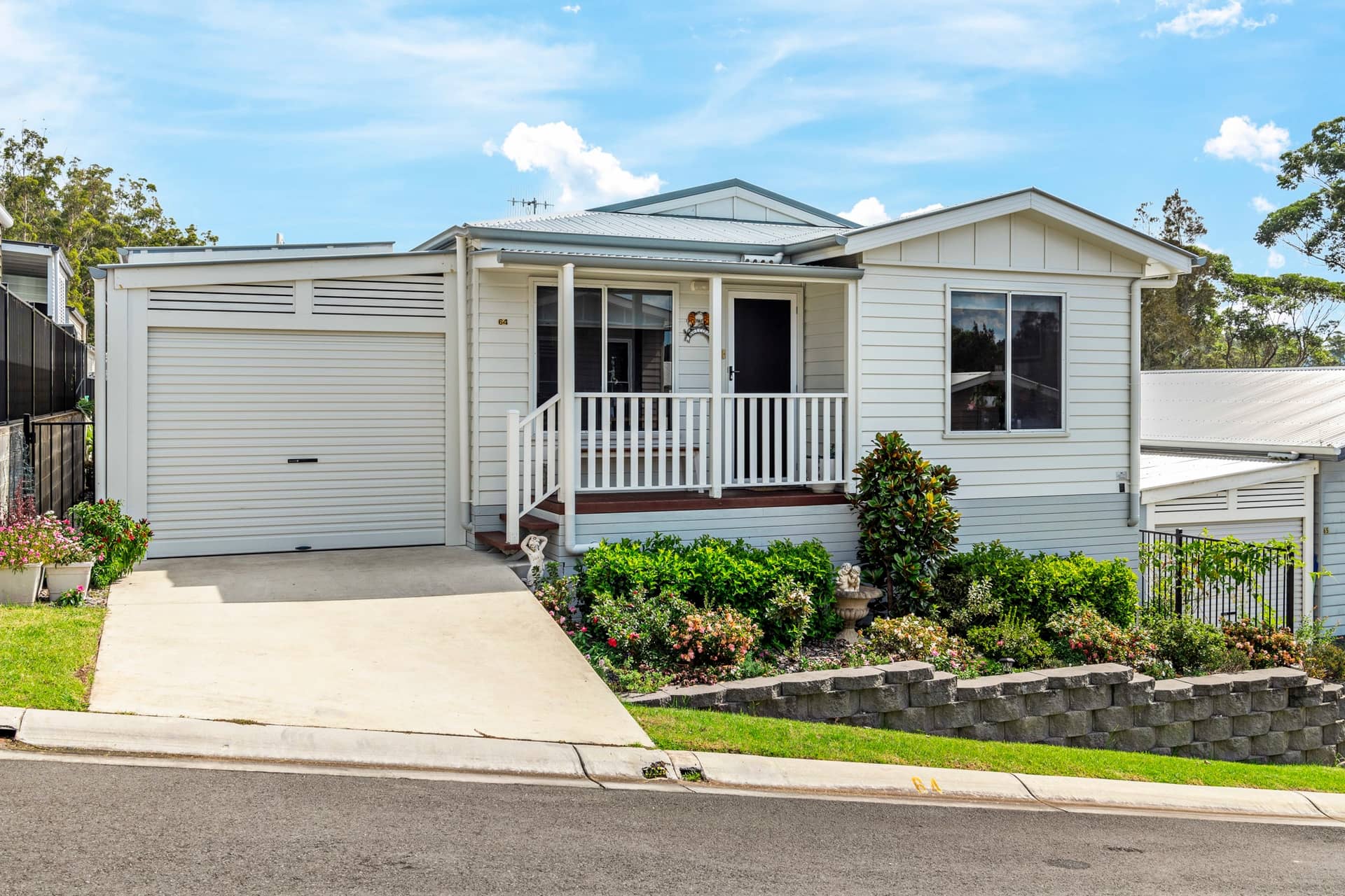 A white land lease home in an Ingenia Lifestyle community with an attached garage, front porch, and garden beds.