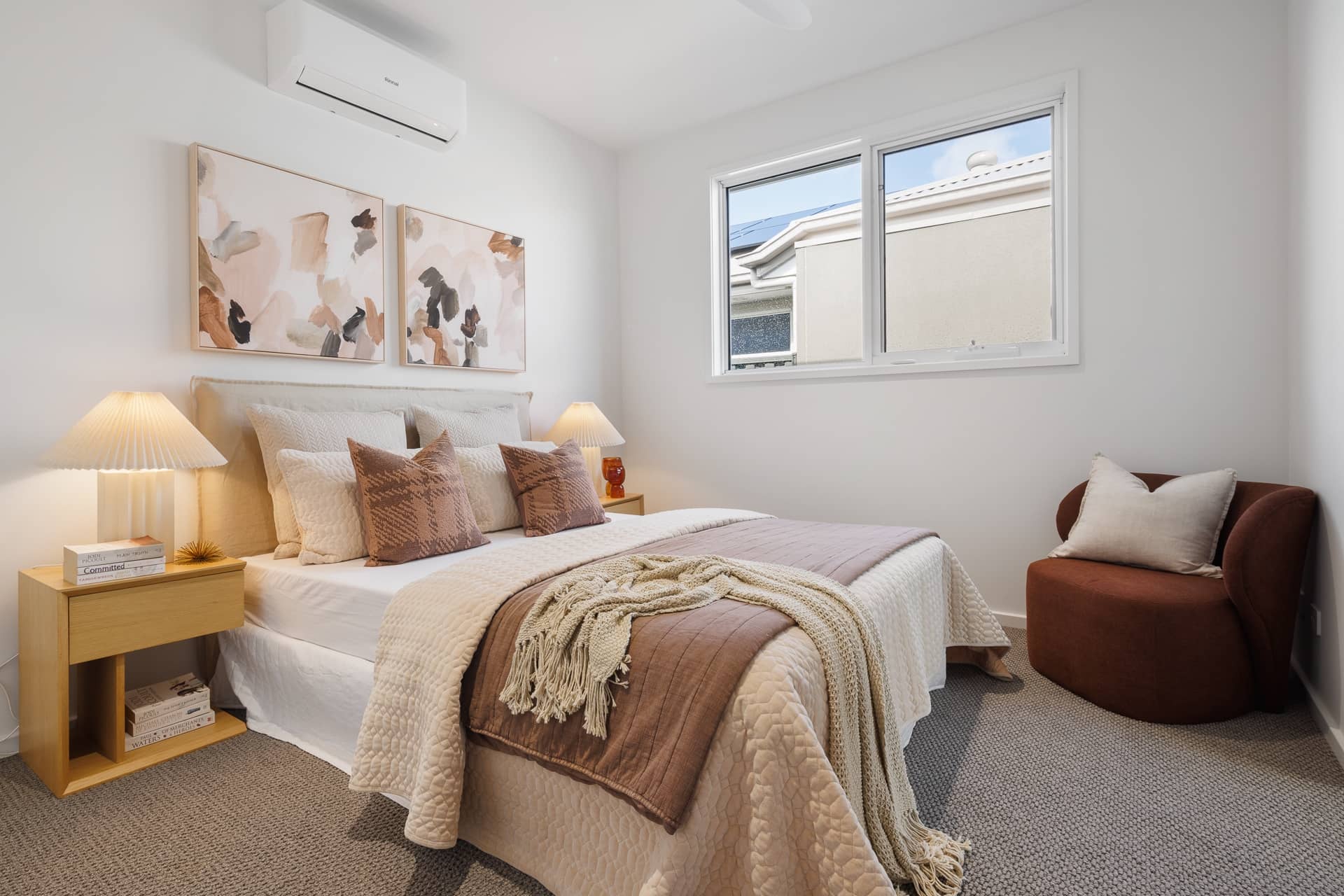 Modern bedroom in an Ingenia Lifestyle home with a bed, side tables, lamps, window, air conditioning, and a brown armchair.
