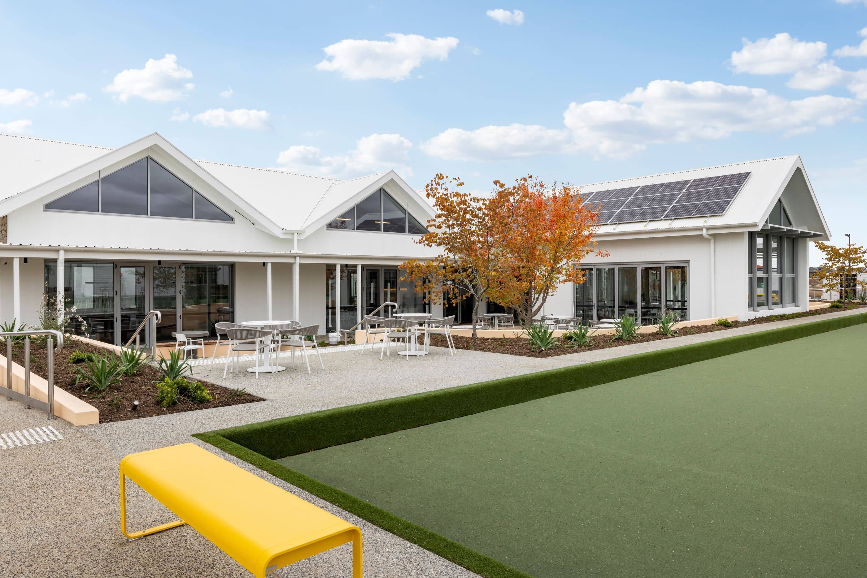 Modern Ingenia Lifestyle community clubhouse with solar panels, outdoor seating, and a green lawn bowls area with a yellow bench.