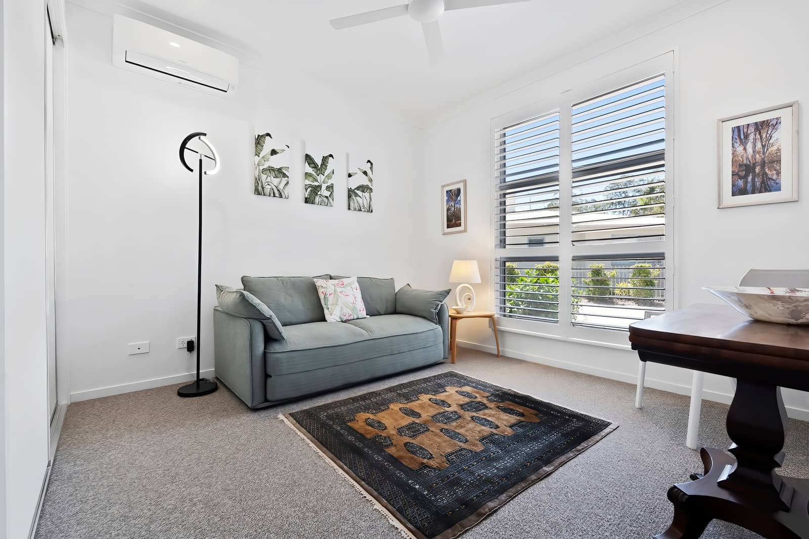 A bright living space in an Ingenia Lifestyle home with a grey sofa, patterned rug, large window with shutters, and air conditioning.