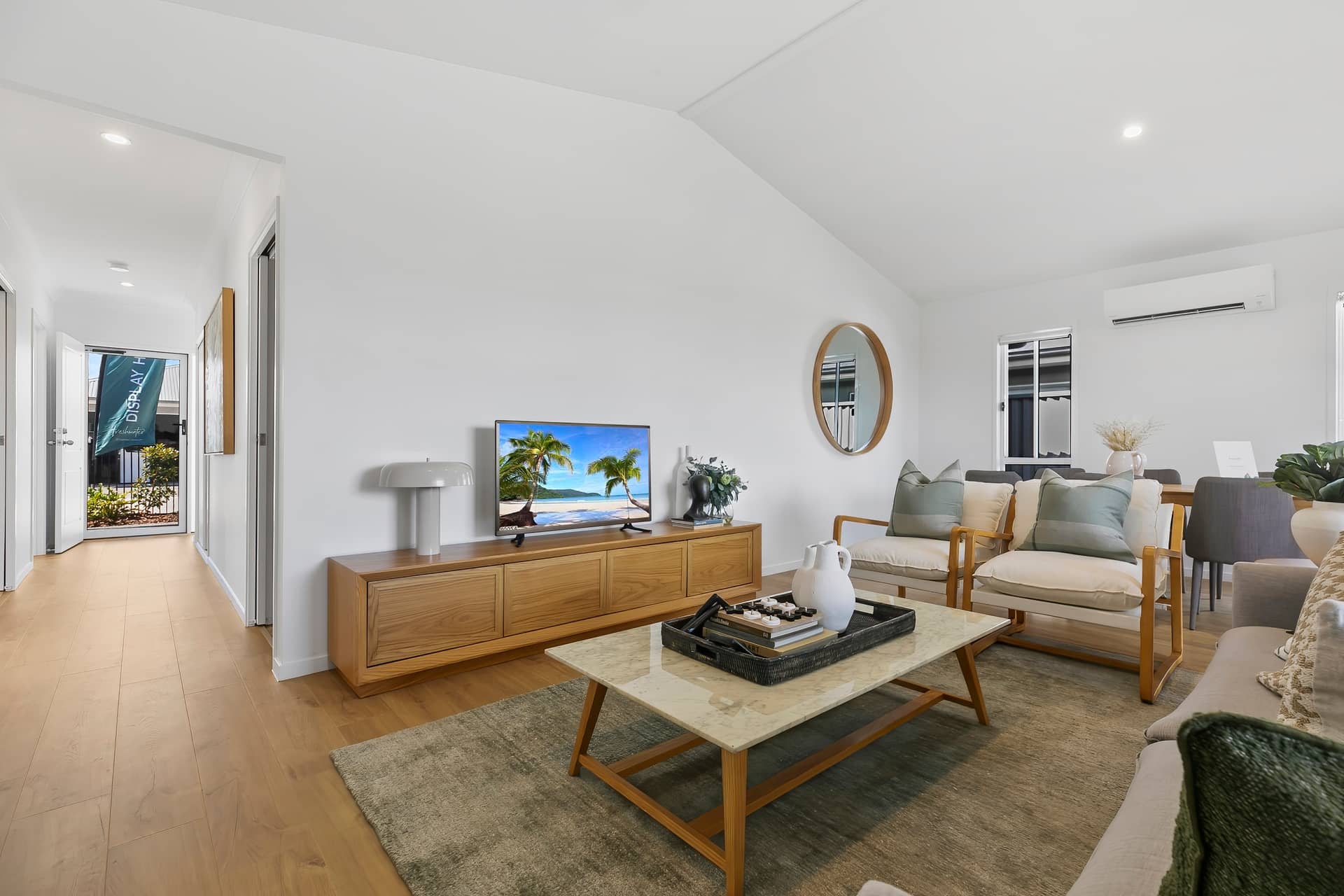 Living room of an Ingenia Lifestyle display home with a vaulted ceiling, timber flooring, TV, sofa, and armchairs.