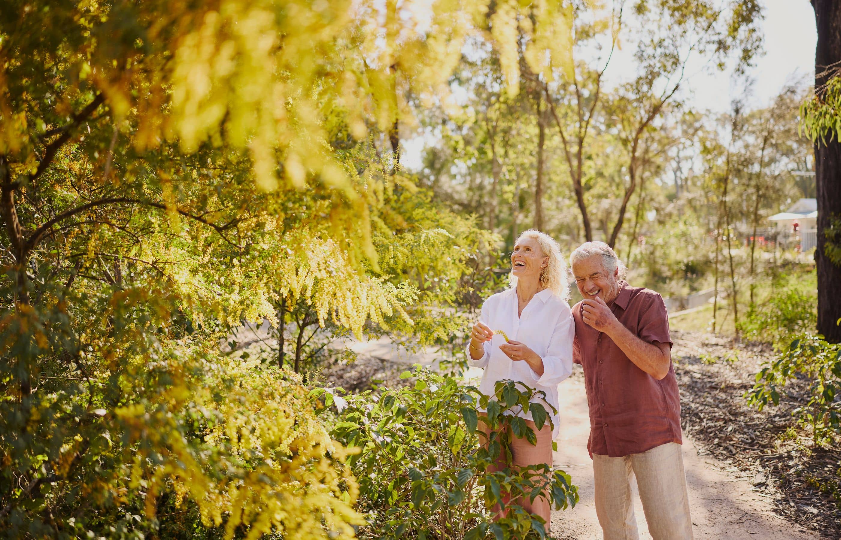 A couple laughs while walking on a path in a sunlit, leafy area of an Ingenia Lifestyle community.