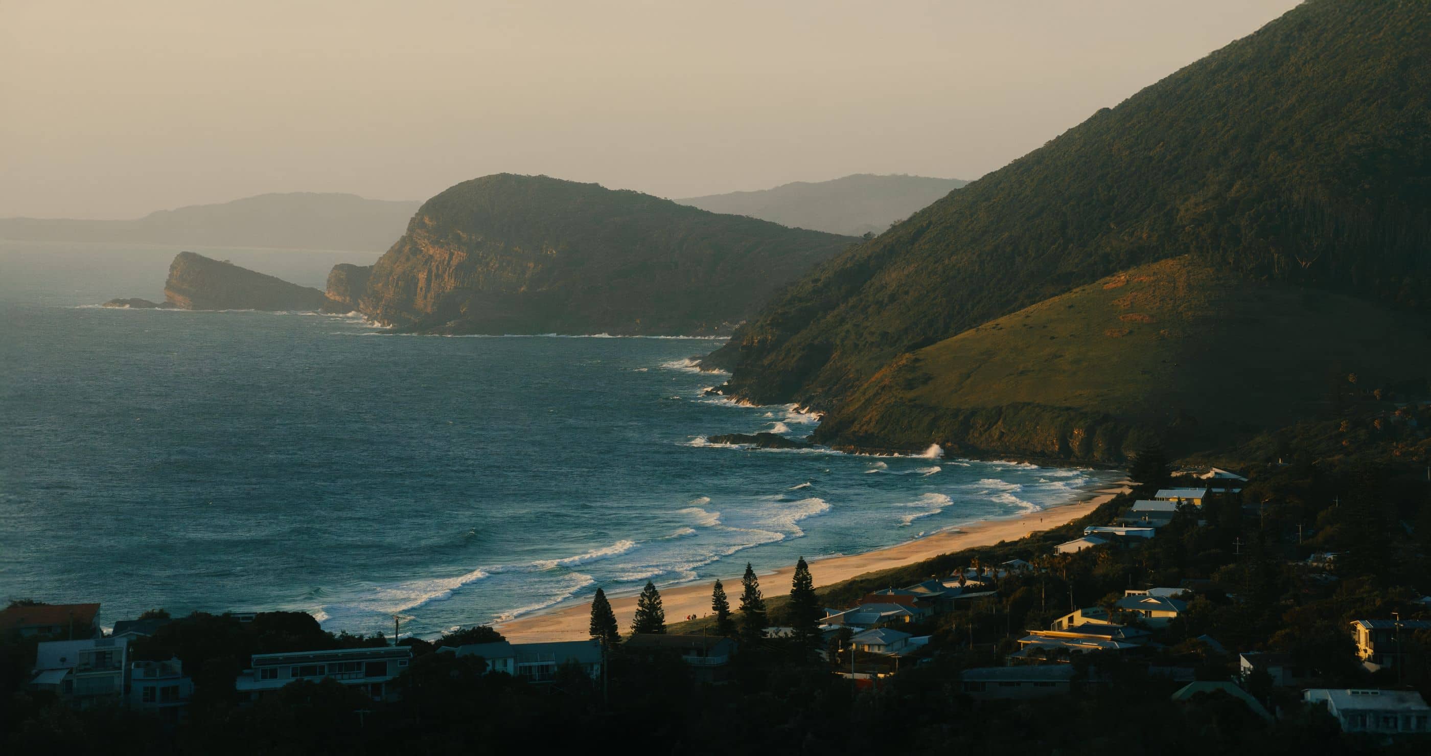 Overhead view of a sandy beach and ocean waves beside green, tree-covered hills with coastal homes.